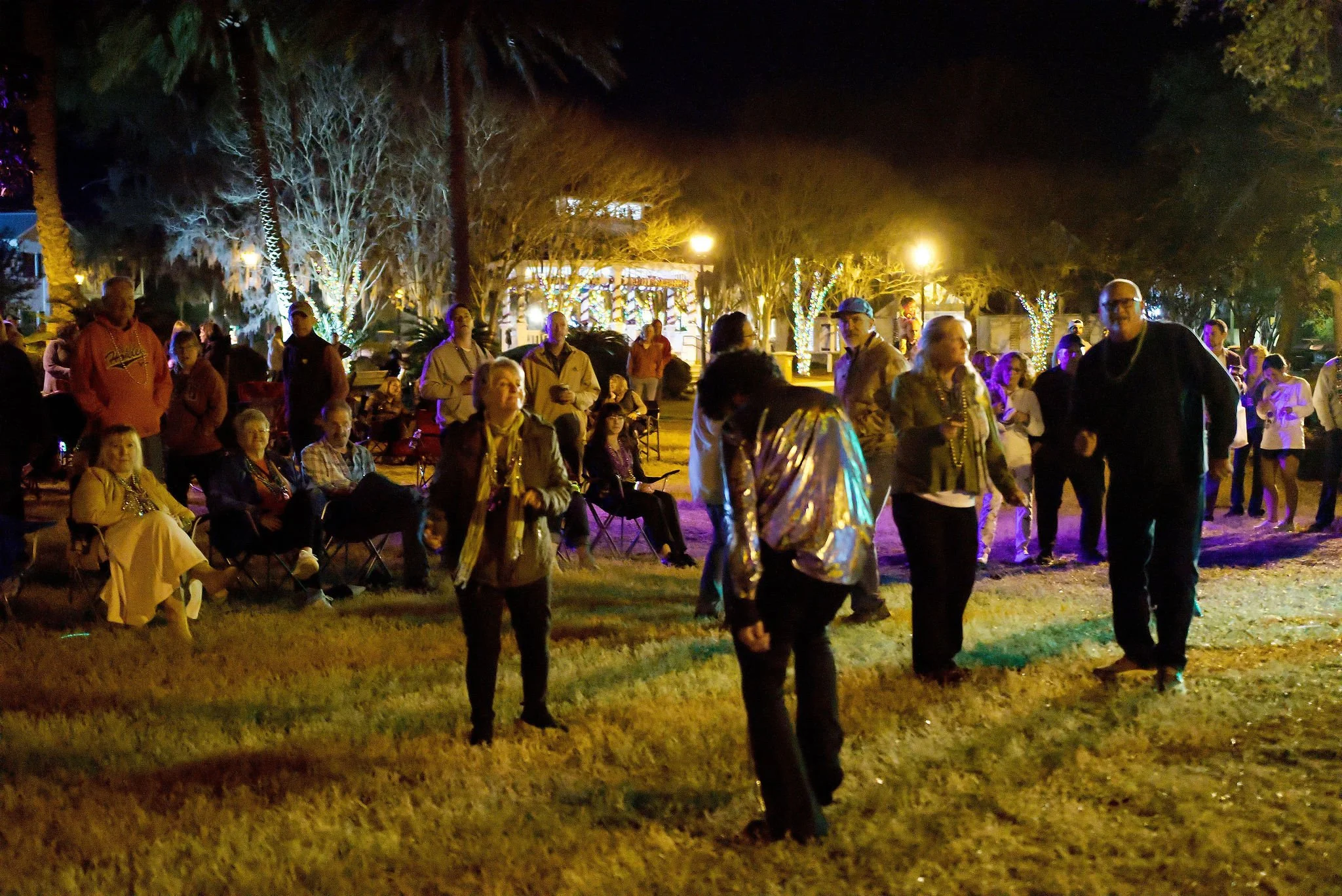 People gathered outdoors at night, some sitting on chairs and others standing, with colorful lights and trees decorated with string lights, suggesting a festive event or outdoor party.
