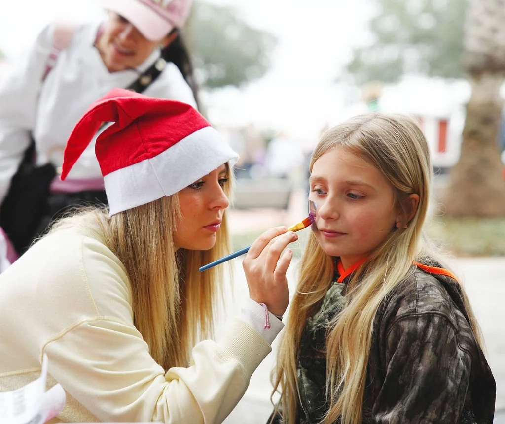 A young girl has her face painted by a woman wearing a Santa hat. The woman is holding a paintbrush and carefully painting on the girl’s face, while another woman in the background, dressed in a white shirt and wearing a Christmas hat, looks on and s