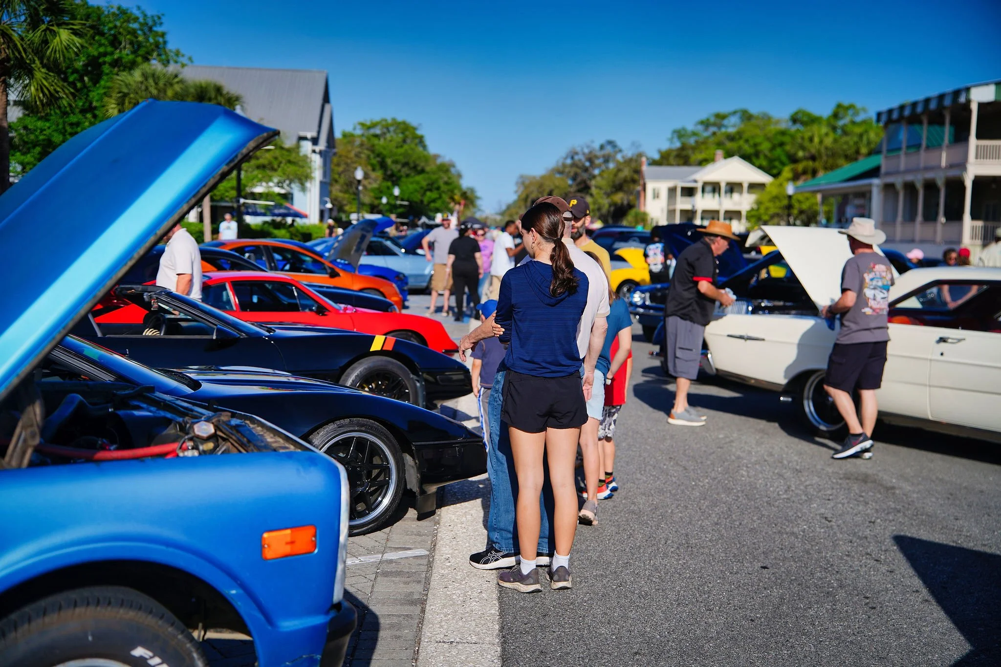 People at a car show viewing classic and sports cars parked with their hoods open on a sunny day, with houses and trees in the background.