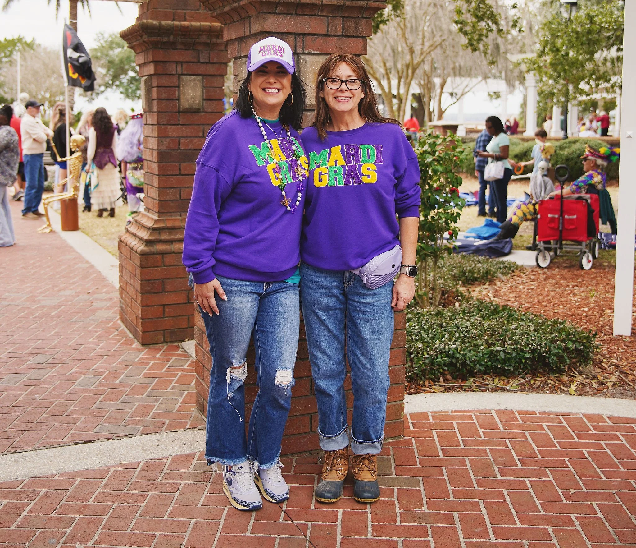 Two women standing close together, smiling, and wearing purple Mardi Gras sweatshirts, in front of a brick pillar during a Mardi Gras celebration with other people, some in costumes, in the background.