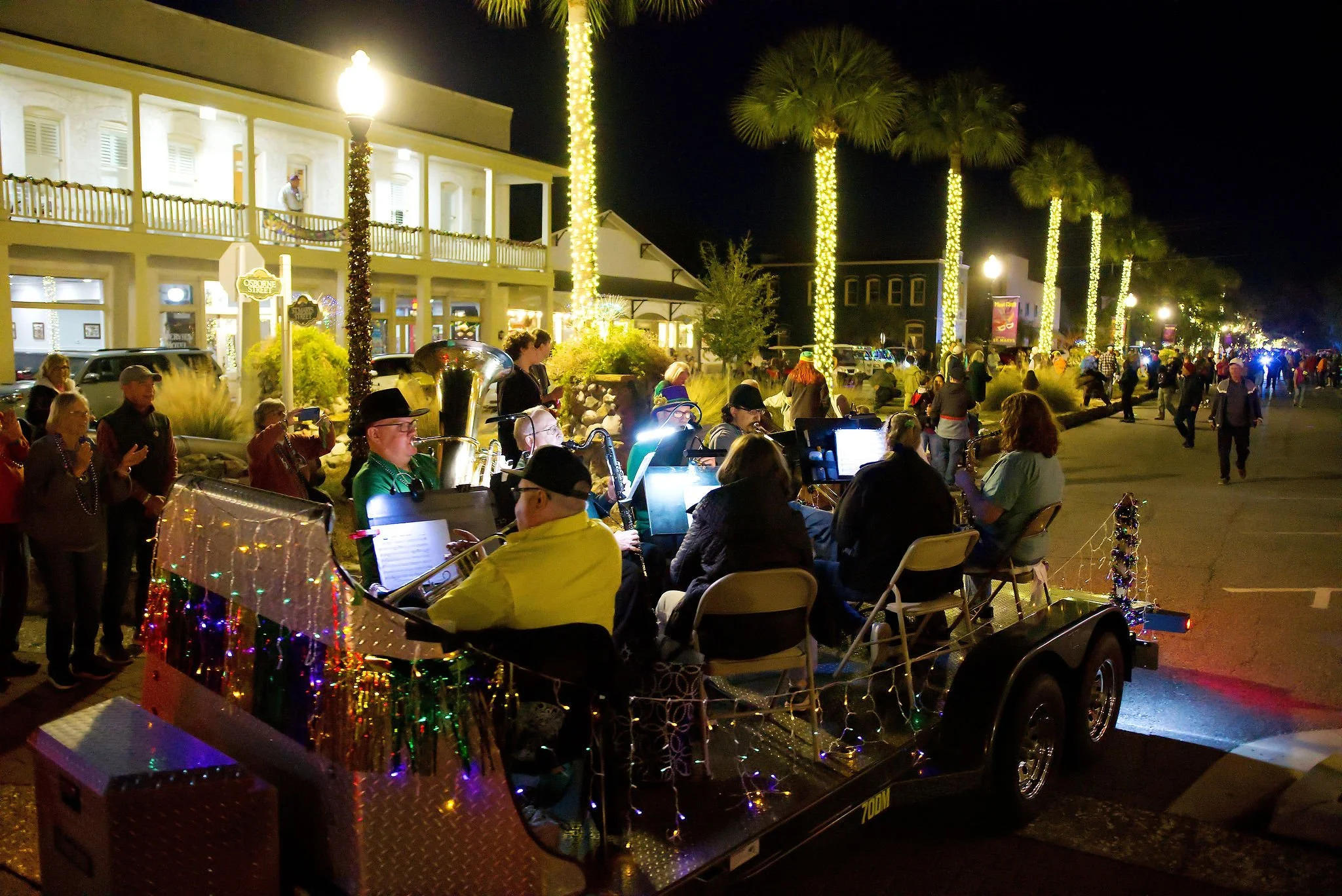 Scene of musicians performing on a trailer decorated with Christmas lights during a night parade, with a crowd of people watching along a decorated street lined with tall, illuminated palm trees.