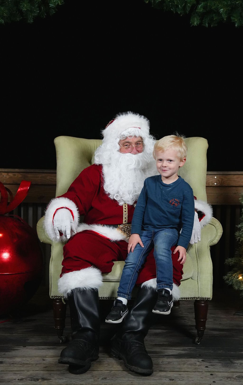 A young boy sitting on Santa Claus's lap during a Christmas photo. Santa is dressed in a traditional red suit with white fur trim, glasses, and a white beard. The setting is festive, with Christmas decorations visible around.