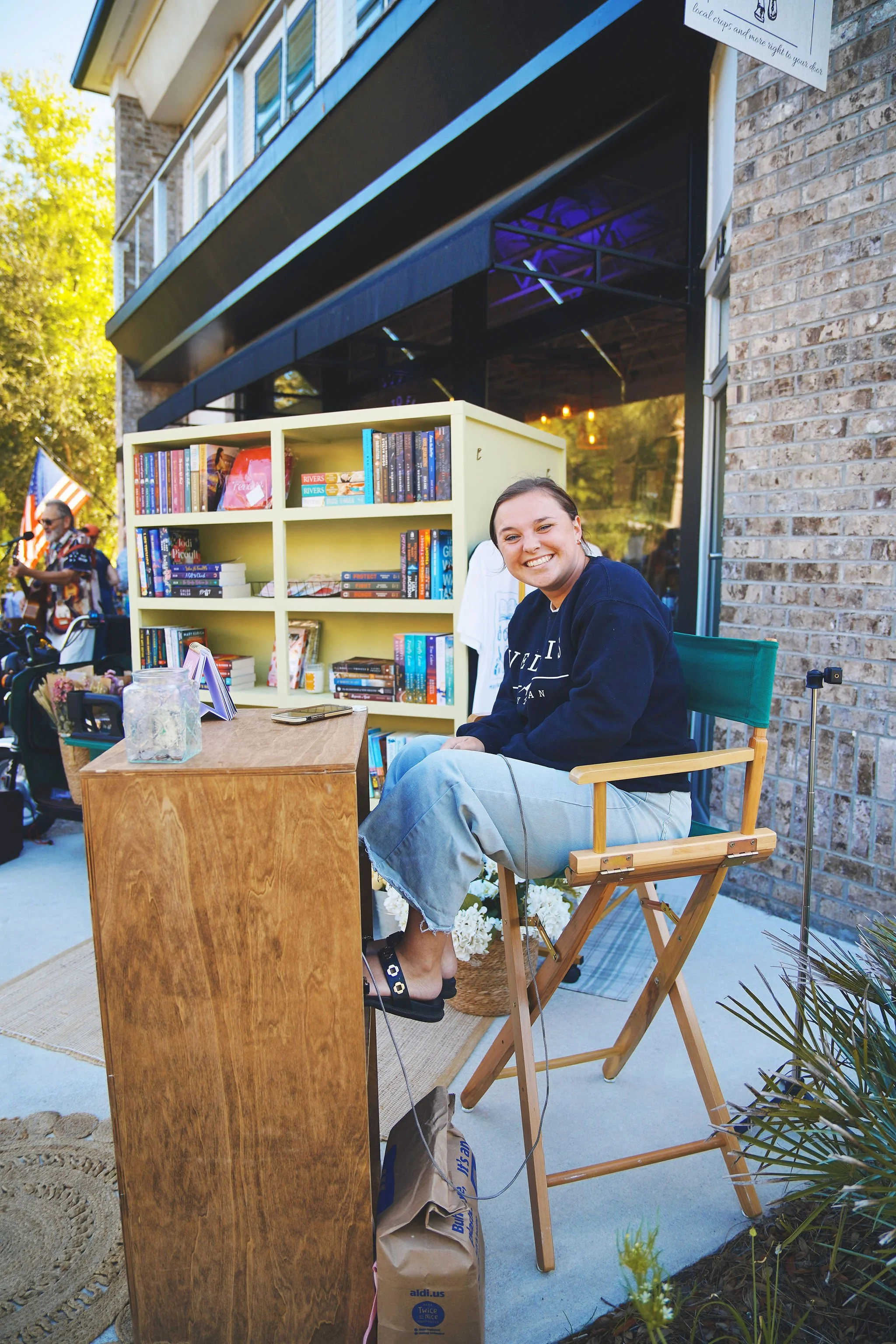 A smiling young woman sitting in a folding chair outdoors at a book sale or signing event, with a bookshelf of colorful books and a wooden table in front of her.