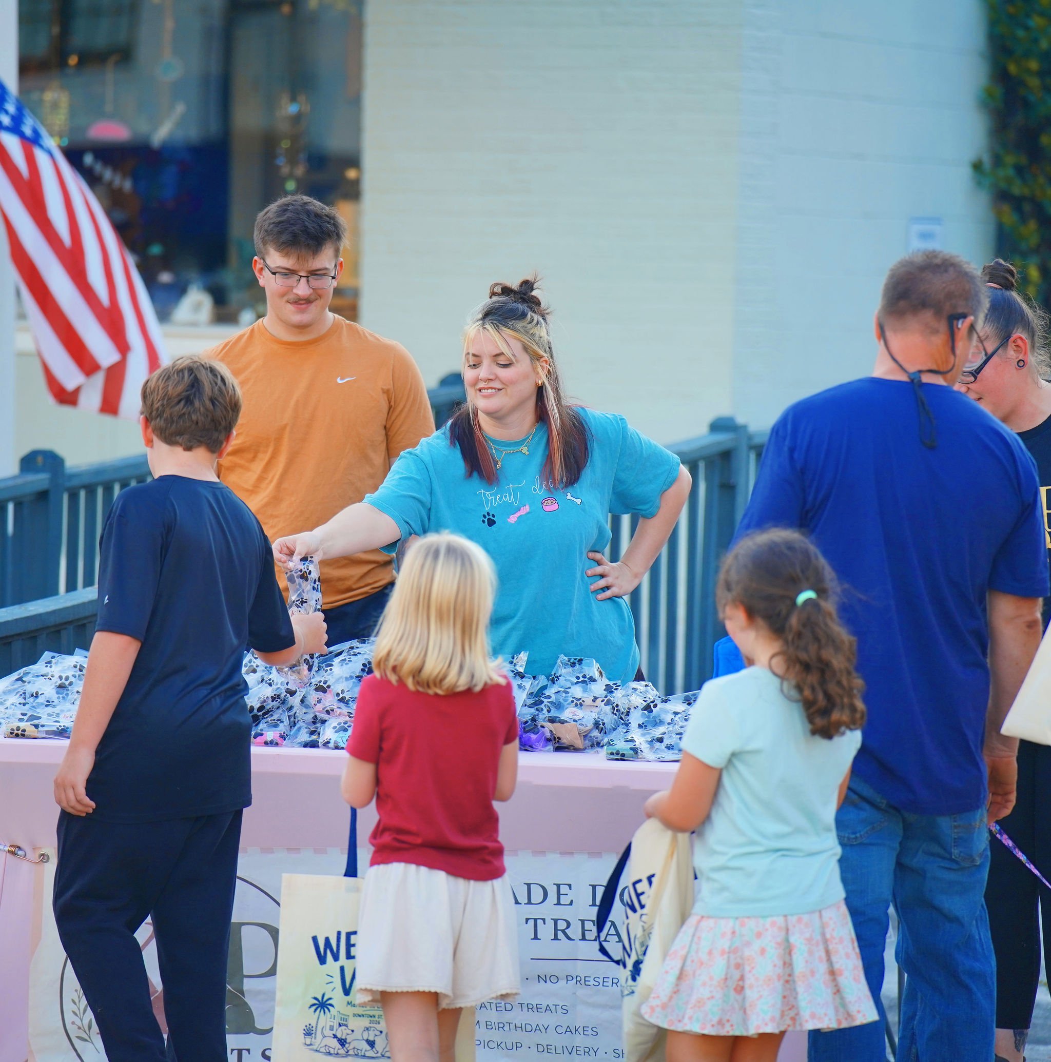 A group of people including teens and children standing outdoors around a table with a gift bag and a sign, engaging in a conversation or activity during daytime.