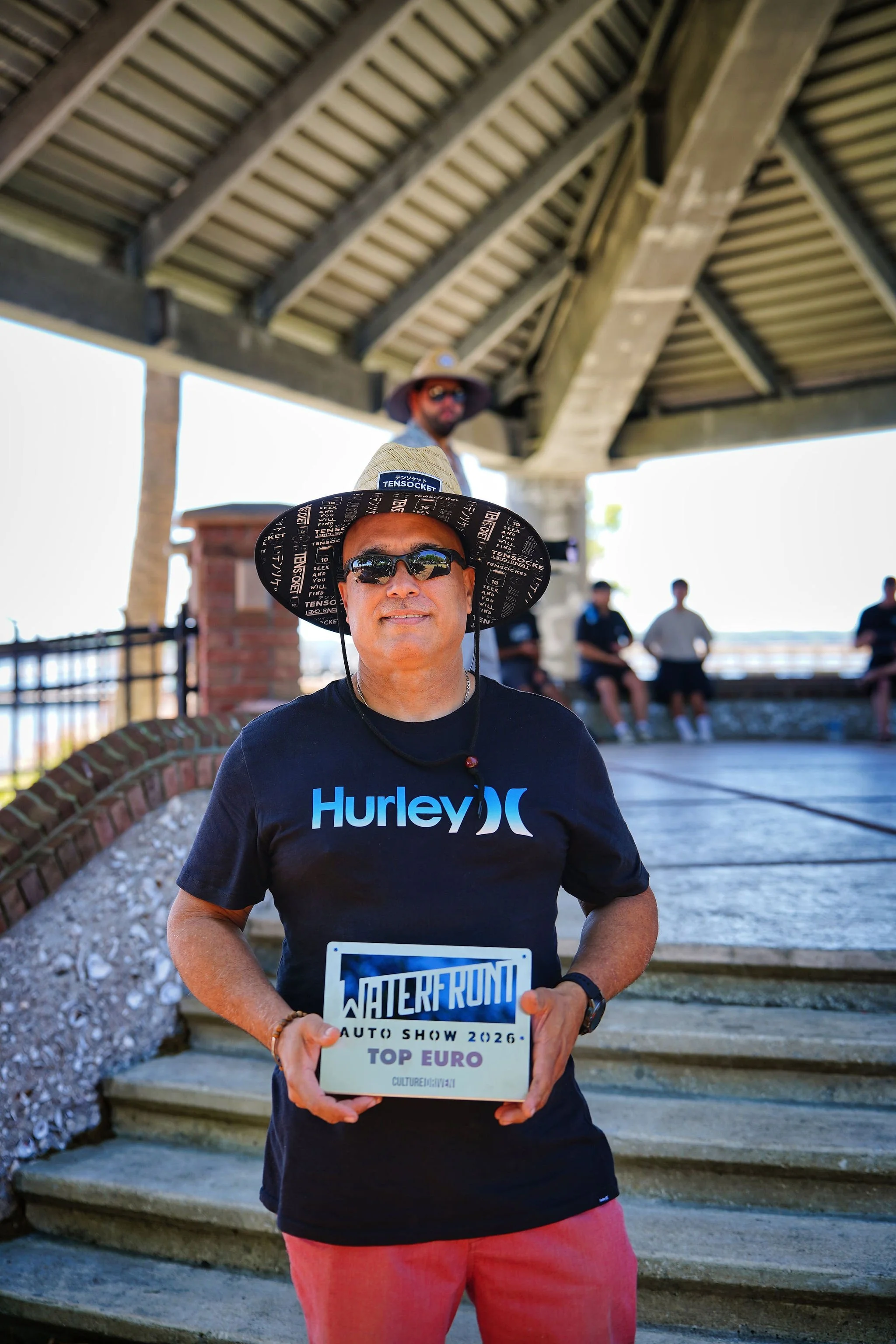 A man wearing sunglasses, a black Hurley shirt, and a wide-brimmed hat is holding a sign that reads "Waterfront Auto Show 2026 Top Euro". He is standing outdoors under a pavilion with stairs and a group of people sitting in the background.