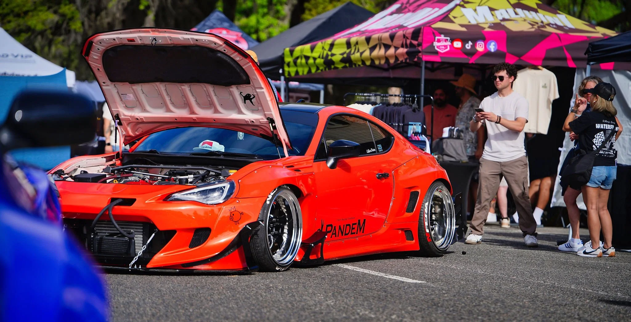 A bright orange modified sports car with its hood open on display at an outdoor event, surrounded by people, tents, and merchandise stalls.