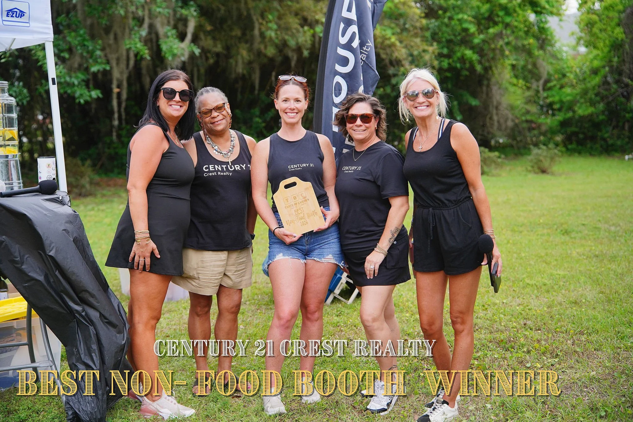 Group of five women standing outdoors at a food booth event. One woman in the center holds a wooden plaque. They are smiling and wearing black clothing. The background includes green trees and a flag with the word "BOUT."