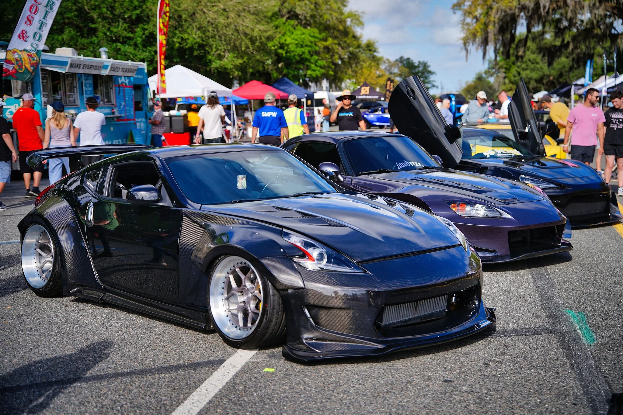 Black sports car with carbon fiber accents at an outdoor car show, with other exotic cars and people in the background.