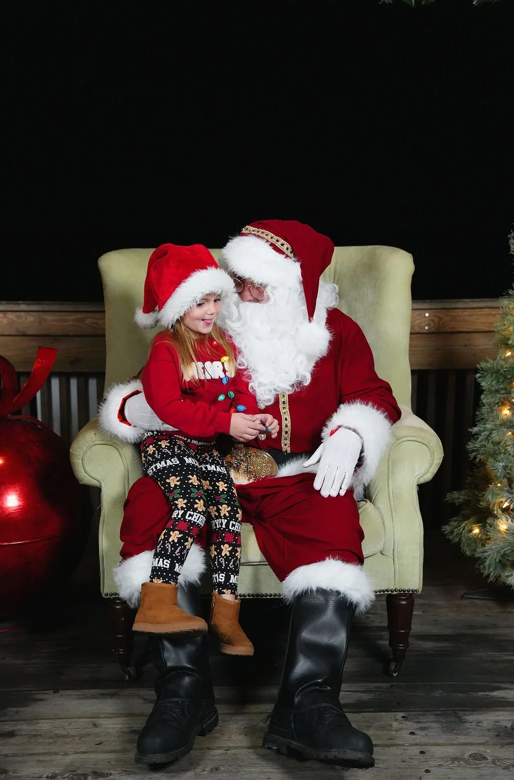 A young girl sitting on Santa Claus's lap, both wearing Christmas hats, in front of a decorated Christmas tree, with a black background and wooden floor.