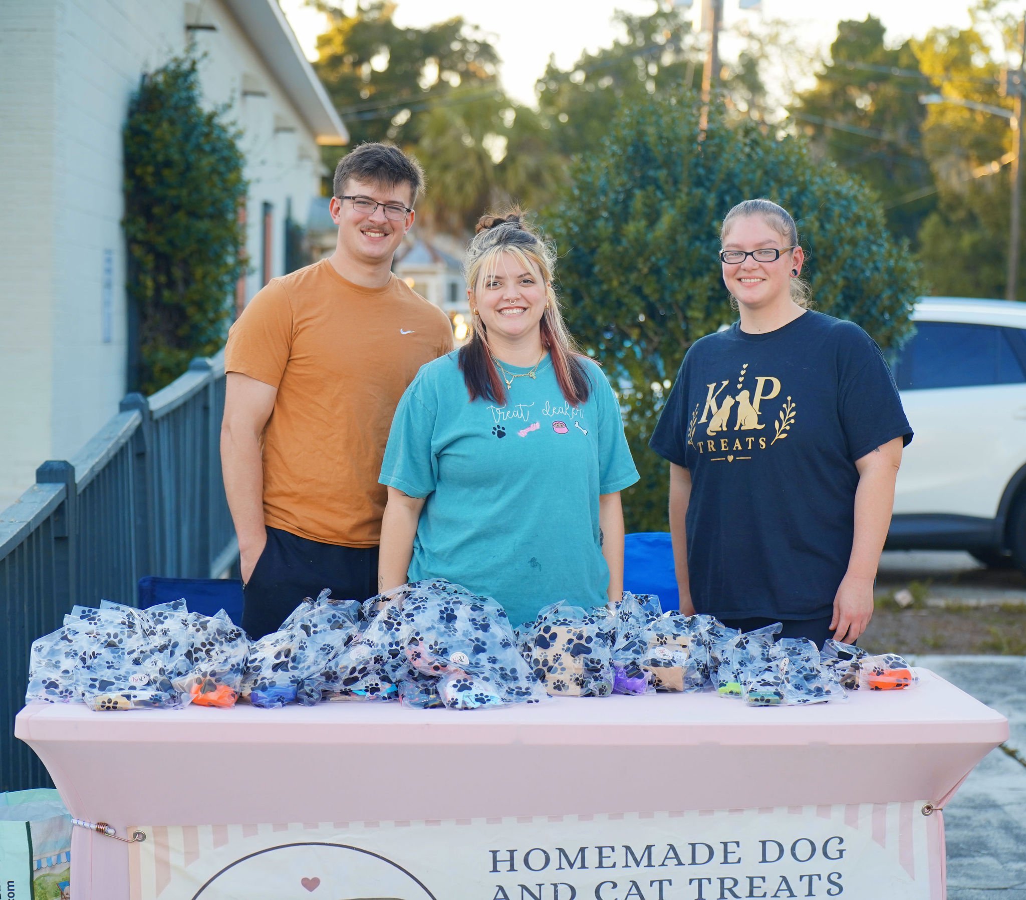 Three people standing behind a table with bags of dog treats, at a stand selling homemade dog and cat treats, outdoors during daytime.