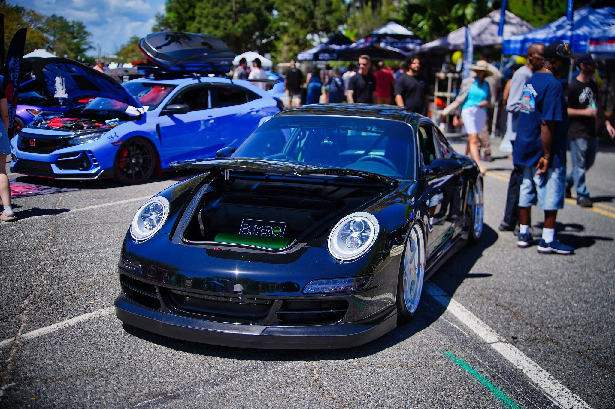 Black sports car with its front trunk open at an outdoor car show, surrounded by people and other cars, including a blue luxury coupe with hood open in the background.