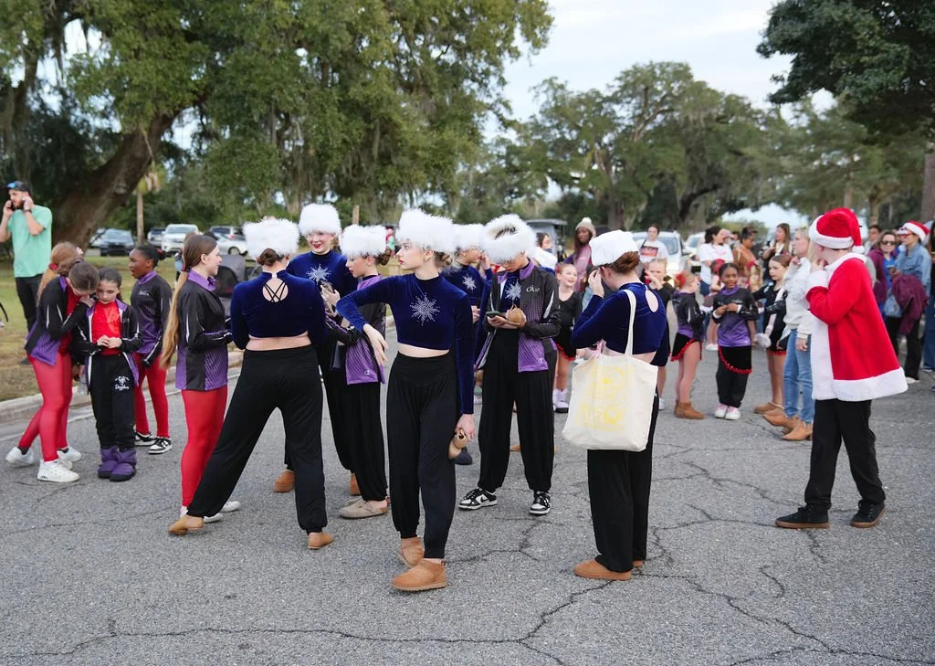 People gathered outdoors for a holiday event, with children and adults, some dressed in Christmas-themed costumes and hats, others in dance or casual attire, under large trees.