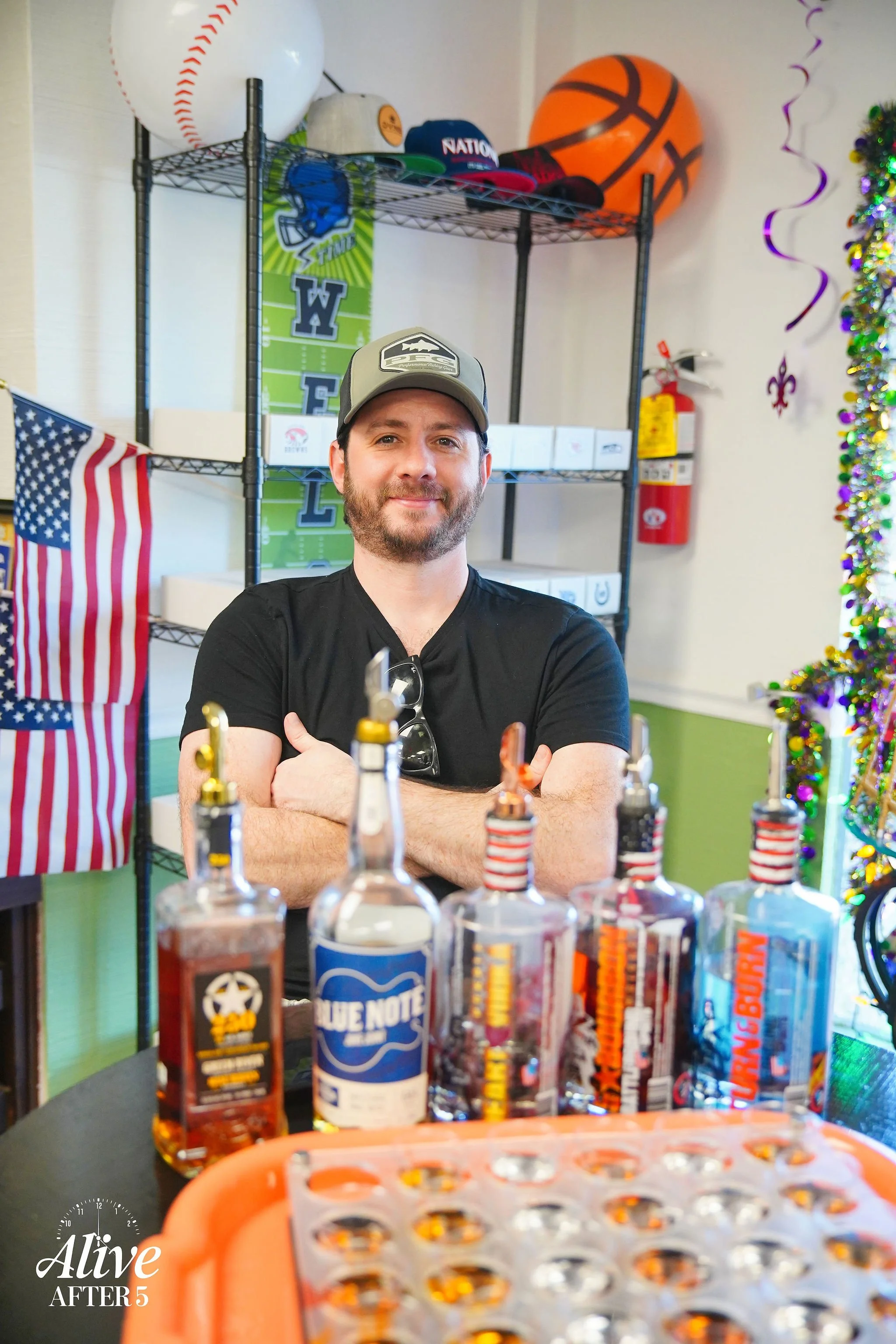 A man with a beard wearing a cap, black t-shirt, and sunglasses hanging from his shirt is standing behind a table displaying various bottles of alcohol and shots. The background includes American flags, a shelf with sports equipment and caps, a fire 