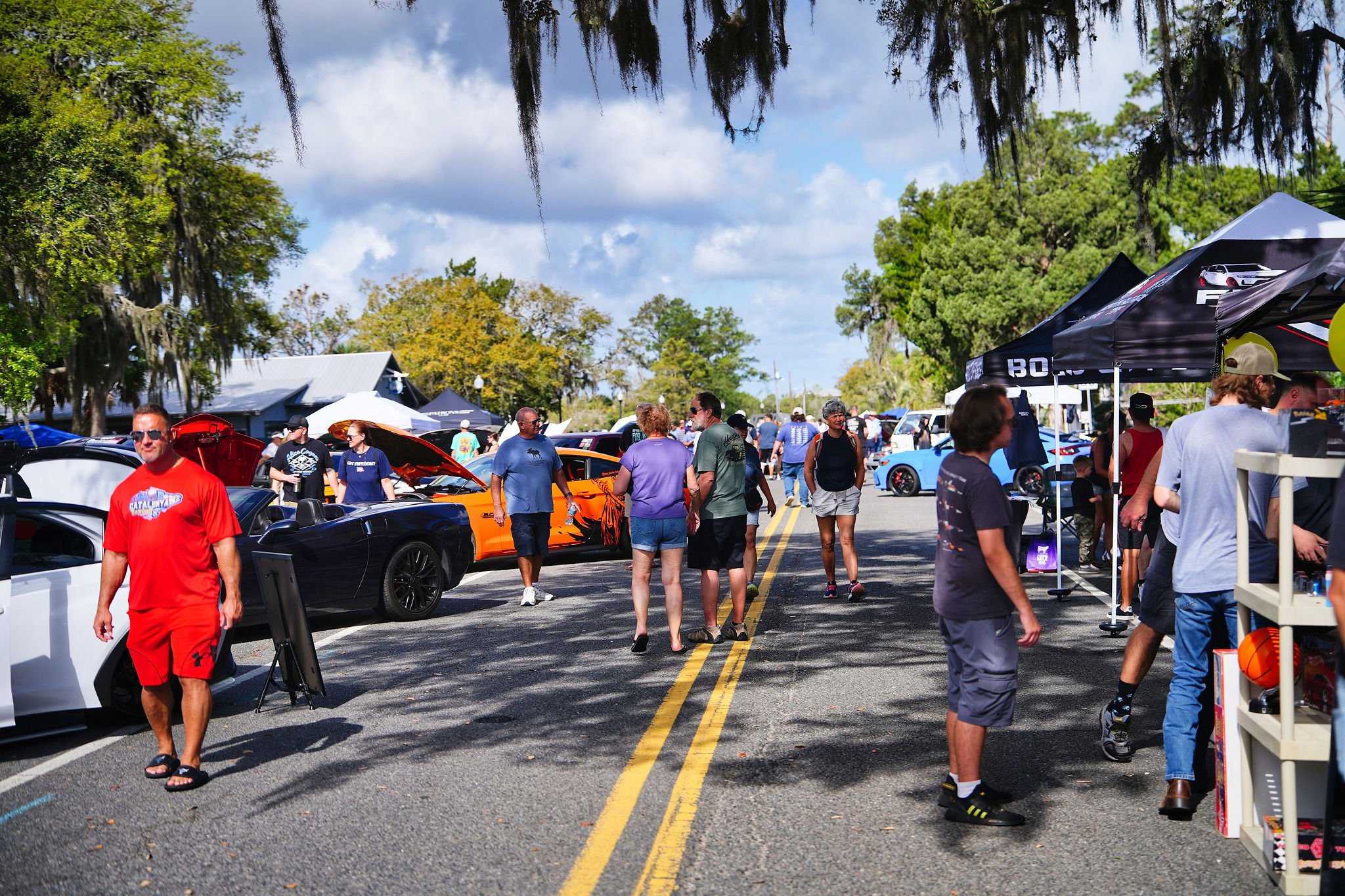 A car show with people walking and browsing parked cars and vendor tents on a sunny day.