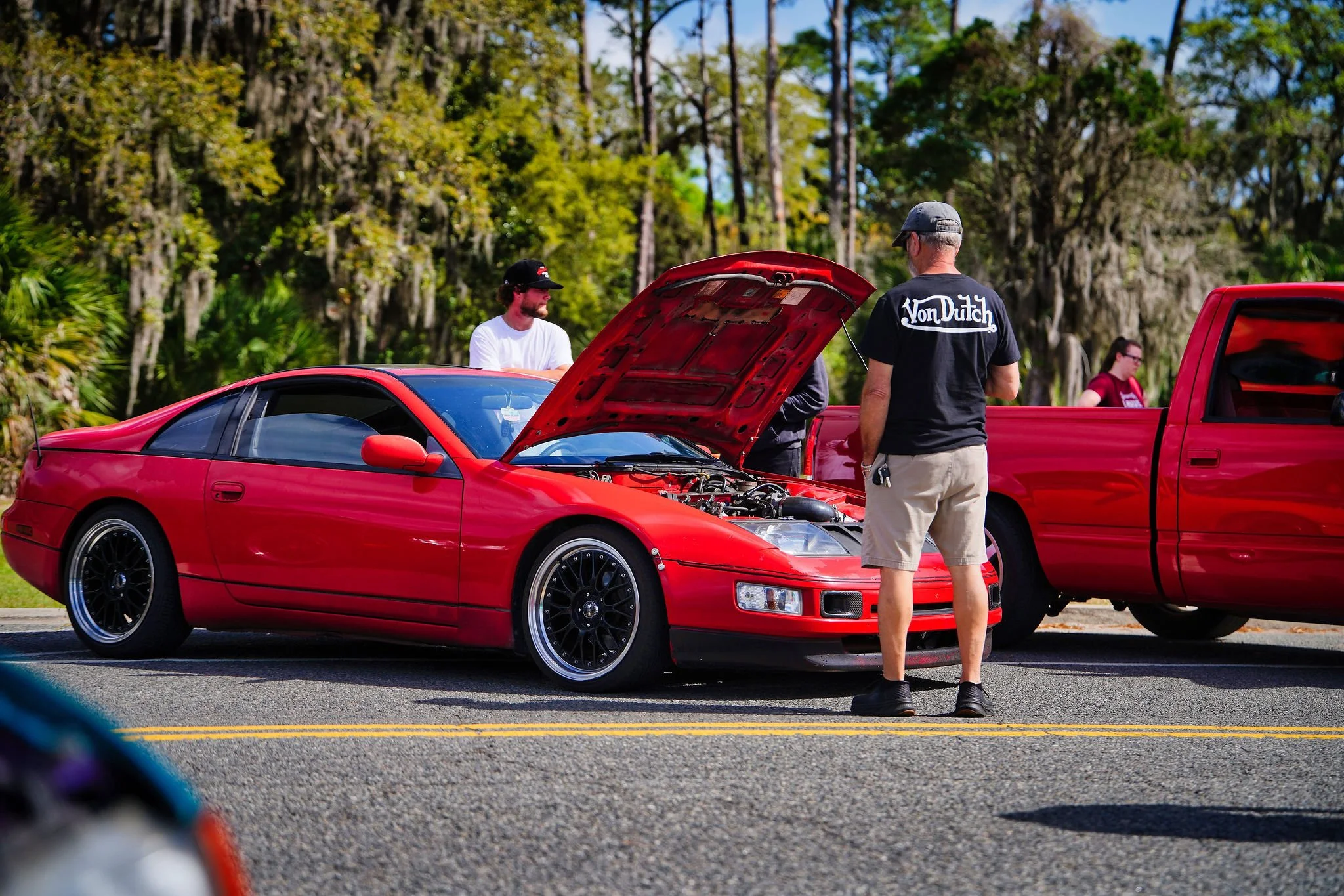 A red sports car with its hood open, parked on a street, surrounded by people. Two men are discussing the car; one facing the camera wearing beige shorts and a black Von Dutch t-shirt, and the other standing behind the car. In the background, two wom
