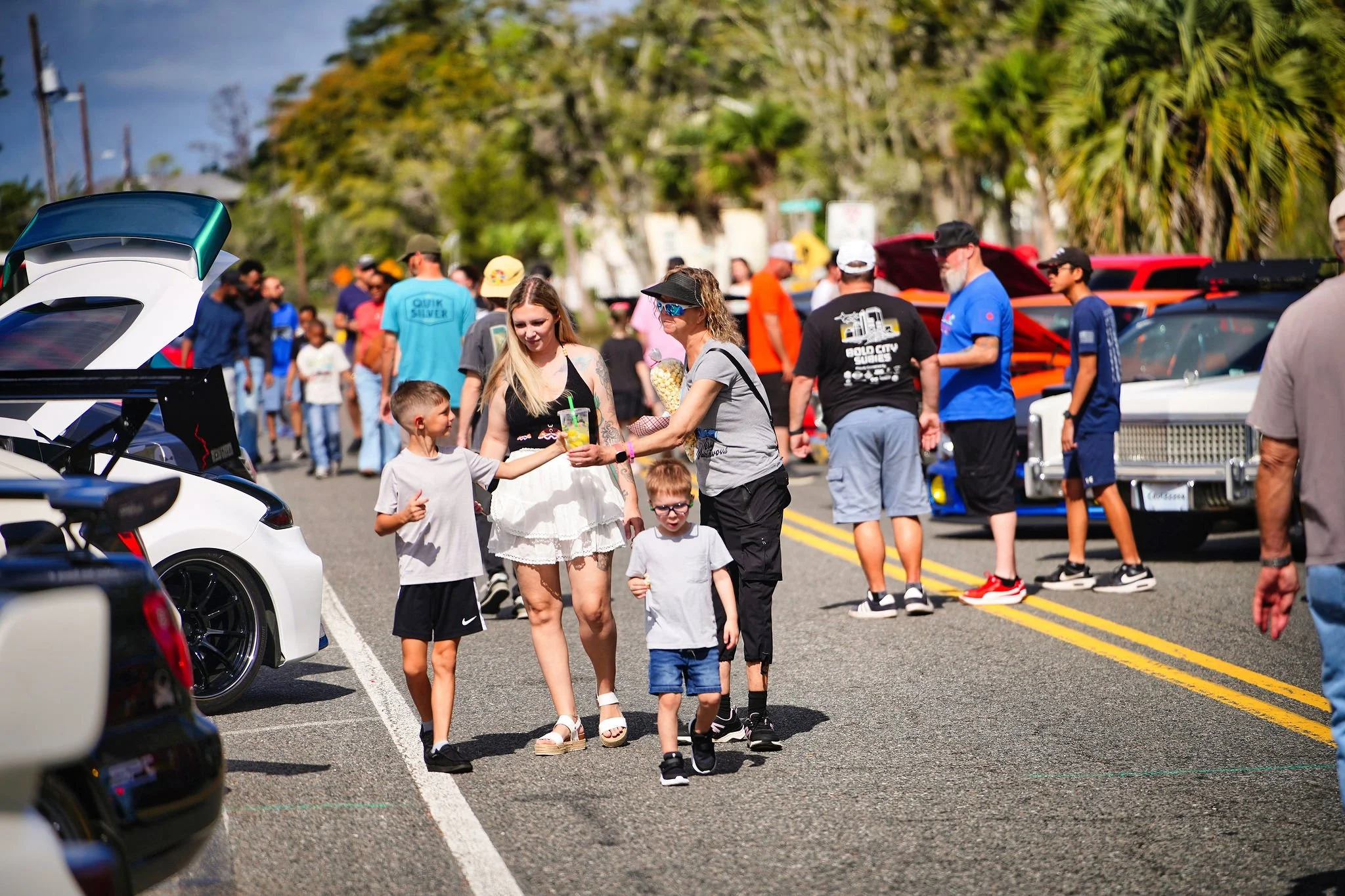 People walking and socializing at a car show on a sunny day, with vintage and modern cars on display along a street lined with trees.