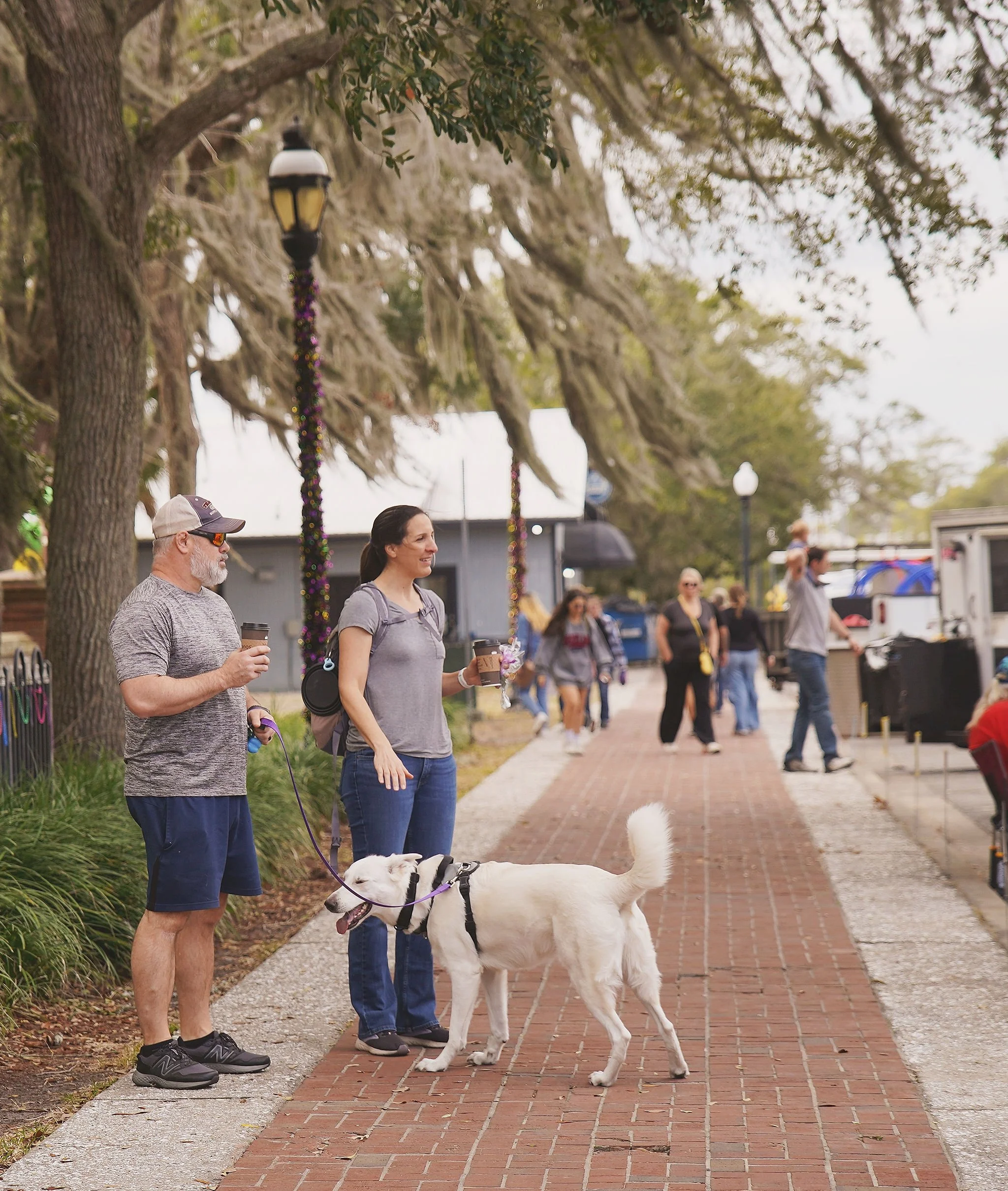 A man and woman standing on a sidewalk with a white dog on a leash, holding cups of coffee, with several people in the background, trees, street lamps, and a food truck on a cloudy day.
