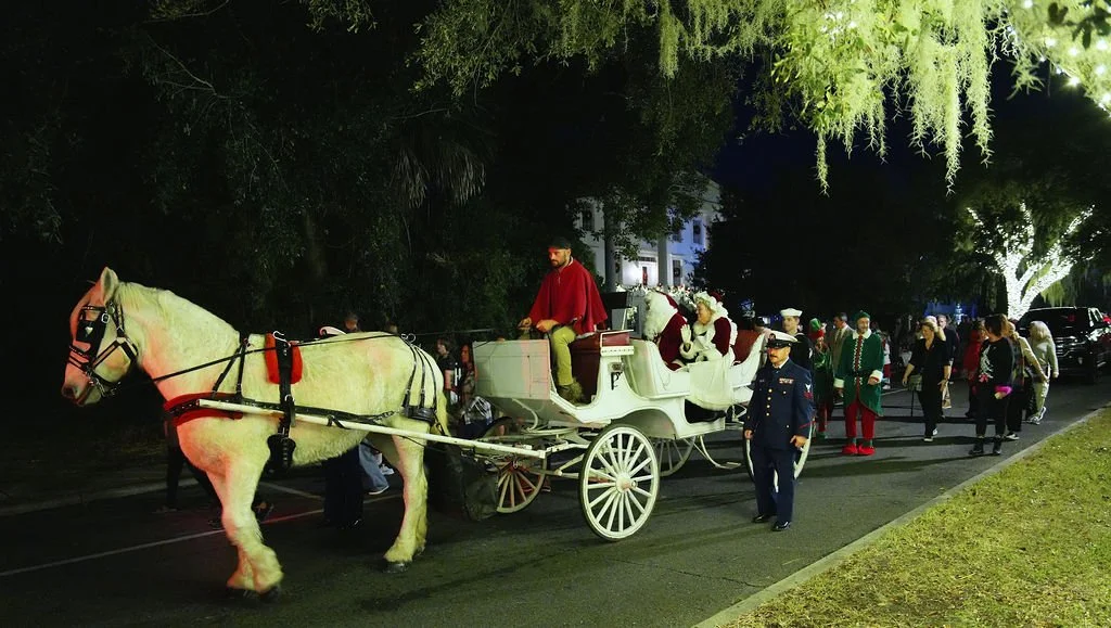A horse-drawn carriage carrying Santa Claus and reindeer figures, followed by people dressed in Christmas costumes, walking on a city street at night.