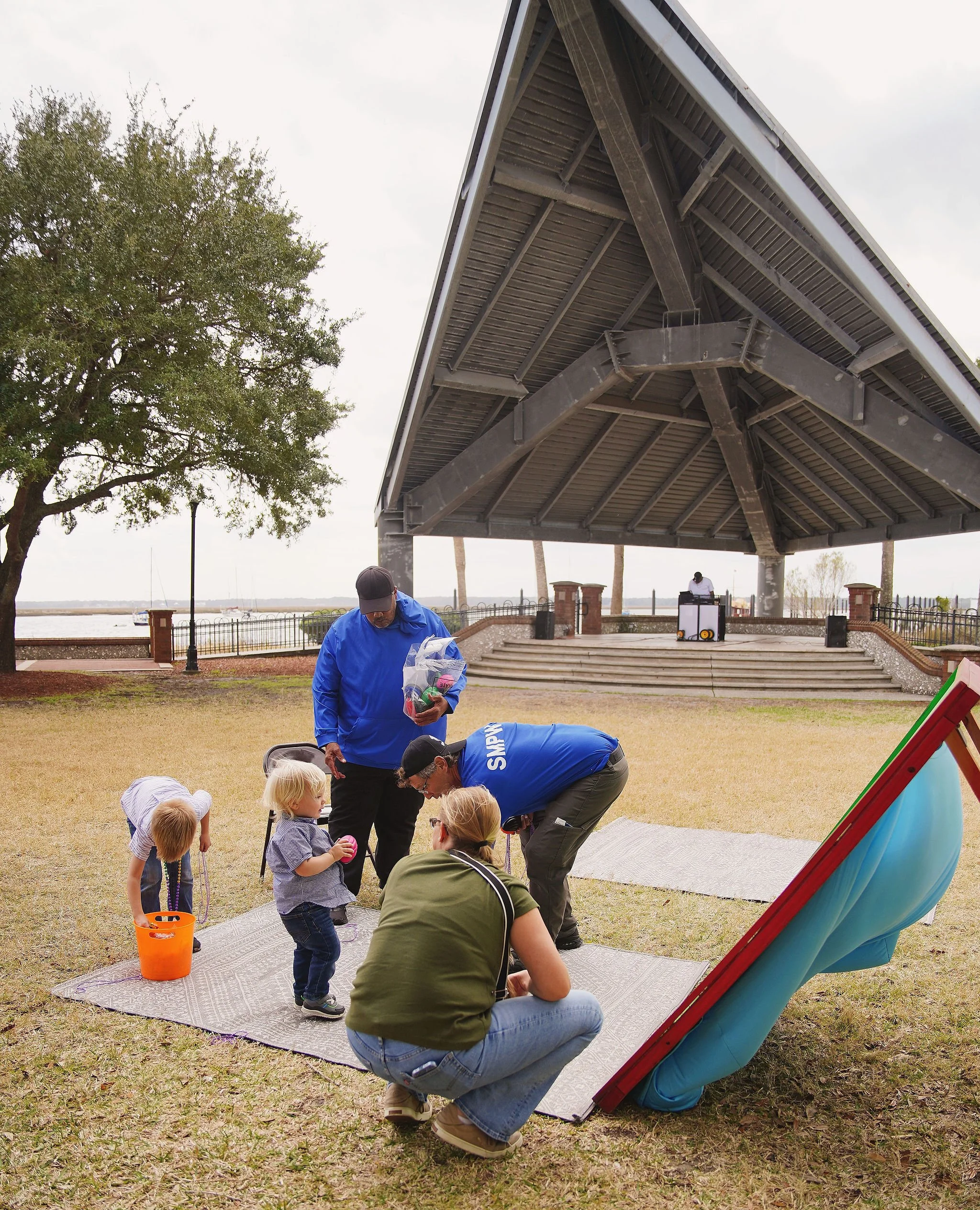 Children and adults participating in an outdoor Easter egg hunt under a park pavilion, with a DJ in the background near a waterfront.
