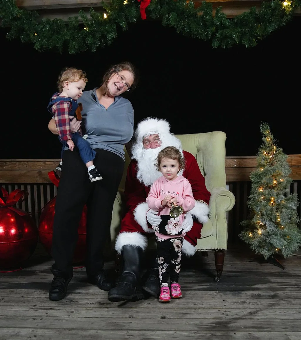 A family with a woman and two young children taking a photo with Santa Claus who is seated in a green armchair. Christmas decorations, including a small decorated tree and large red ornaments, are in the background and at the top of the image.