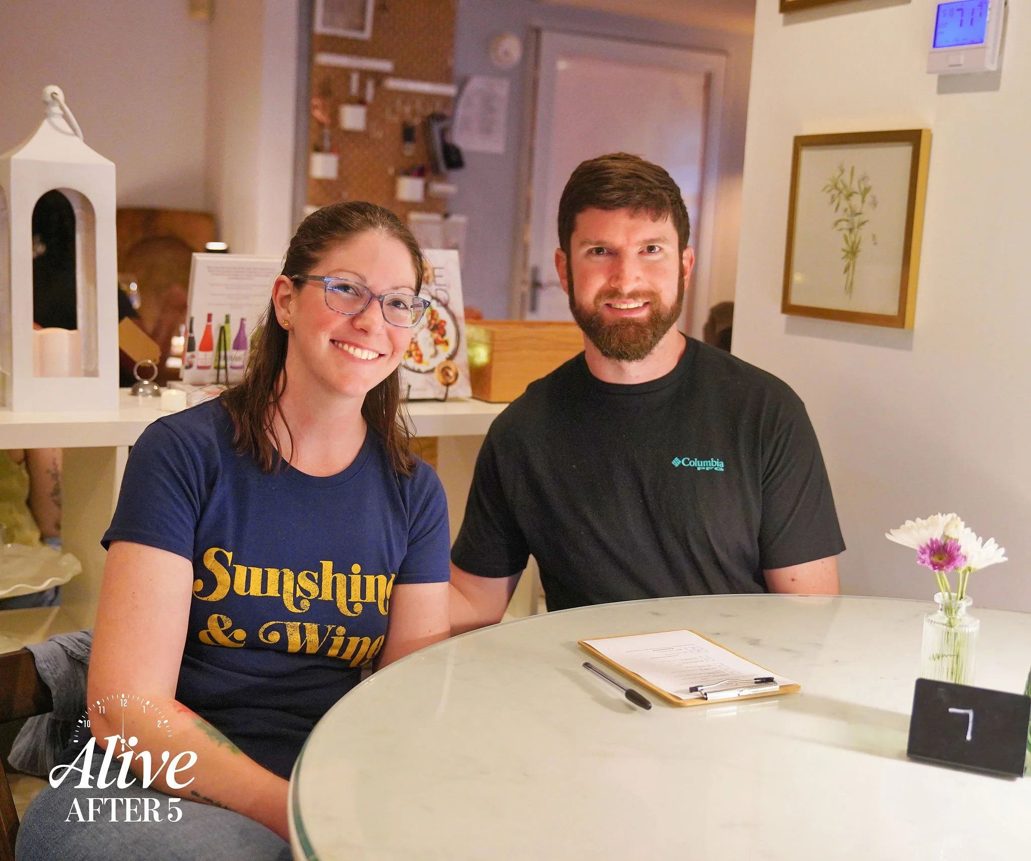 A smiling woman with glasses and a man with a beard sitting at a white marble table in a cozy indoor setting. The woman is wearing a printed navy blue t-shirt, and the man is wearing a black Columbia t-shirt. There is a small vase with pink and white