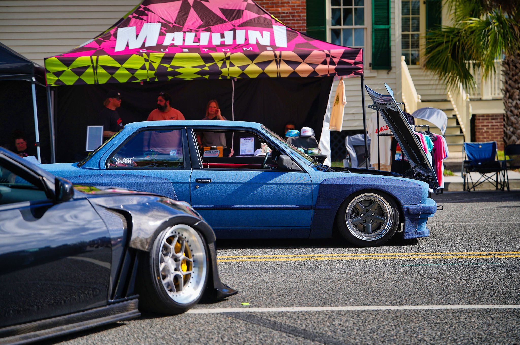A blue modified sports car with lowered suspension at a car show, with its hood open, parked on street in front of a vendor tent.