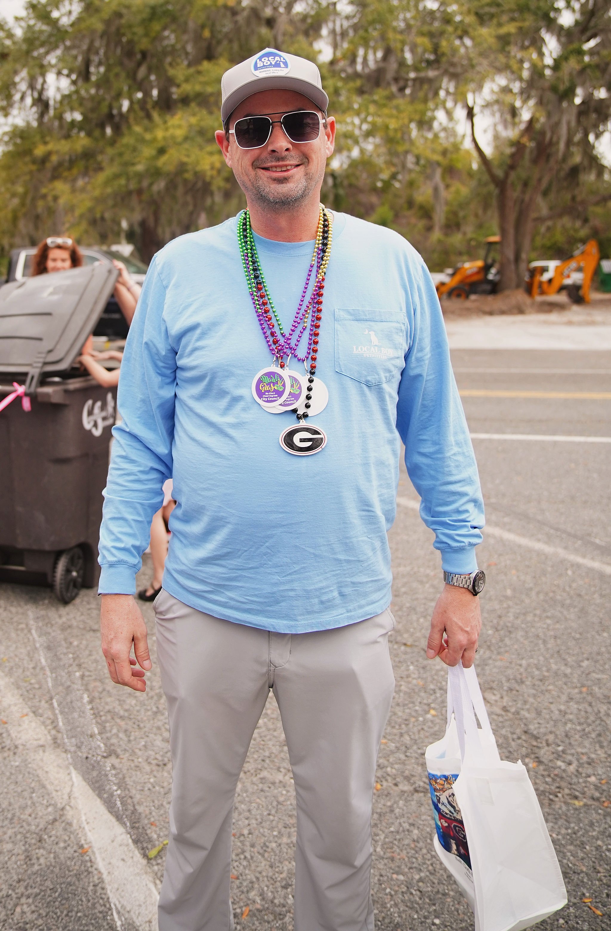 A man wearing sunglasses, a gray baseball cap, a light blue long-sleeve shirt, and beige pants stands outdoors. He is smiling and has several beaded necklaces with various logos and designs around his neck. He is holding a white tote bag in his right