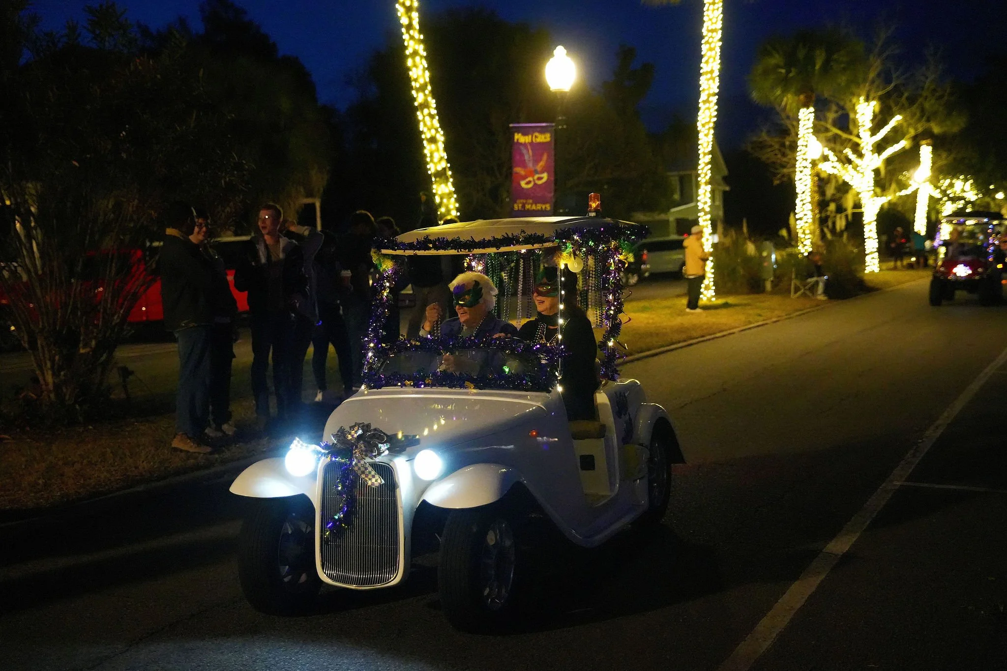 A small white vintage-style car decorated with purple tinsel and bows, with two people inside wearing masks, driving in a parade at night. The street is adorned with Christmas lights on trees, and people are gathered to watch the parade.