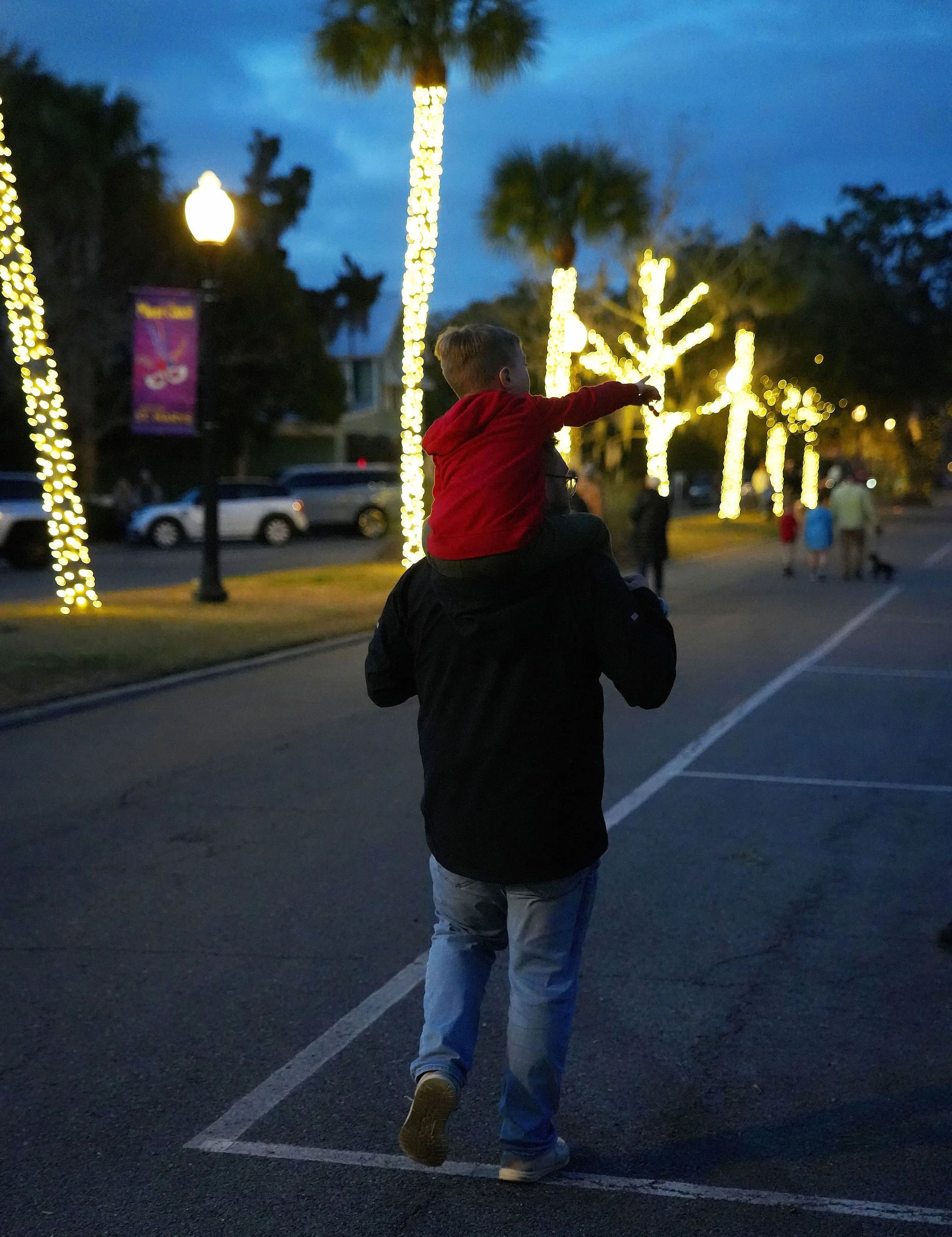 A man carrying a boy on his shoulders walking along a street decorated with Christmas lights. The boy, wearing a red hoodie, points ahead. The scene is set during dusk or evening.