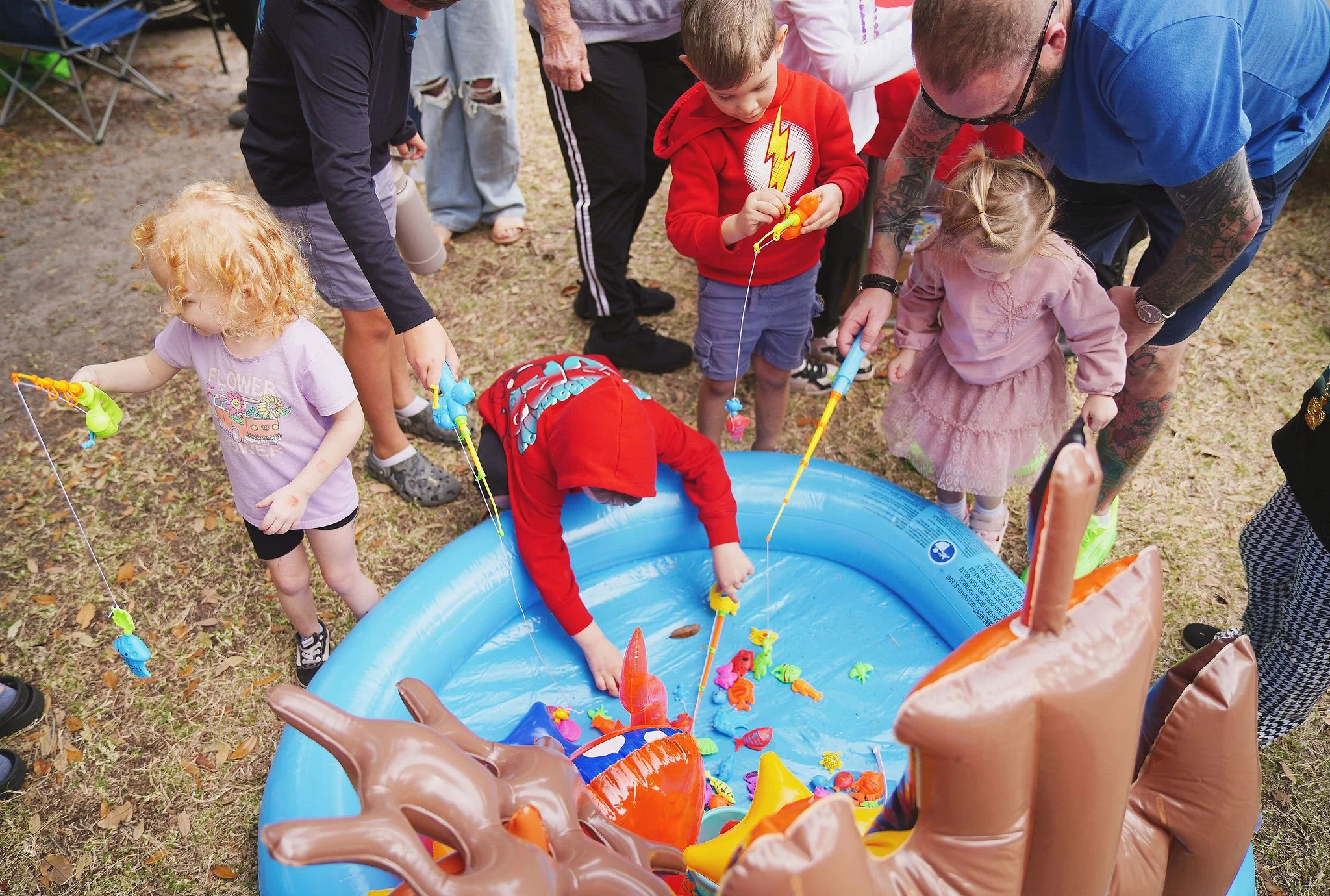 Children and adults playing a game of fishing with toy fishing poles into a small inflatable pool filled with colorful plastic fish and toys at an outdoor event.