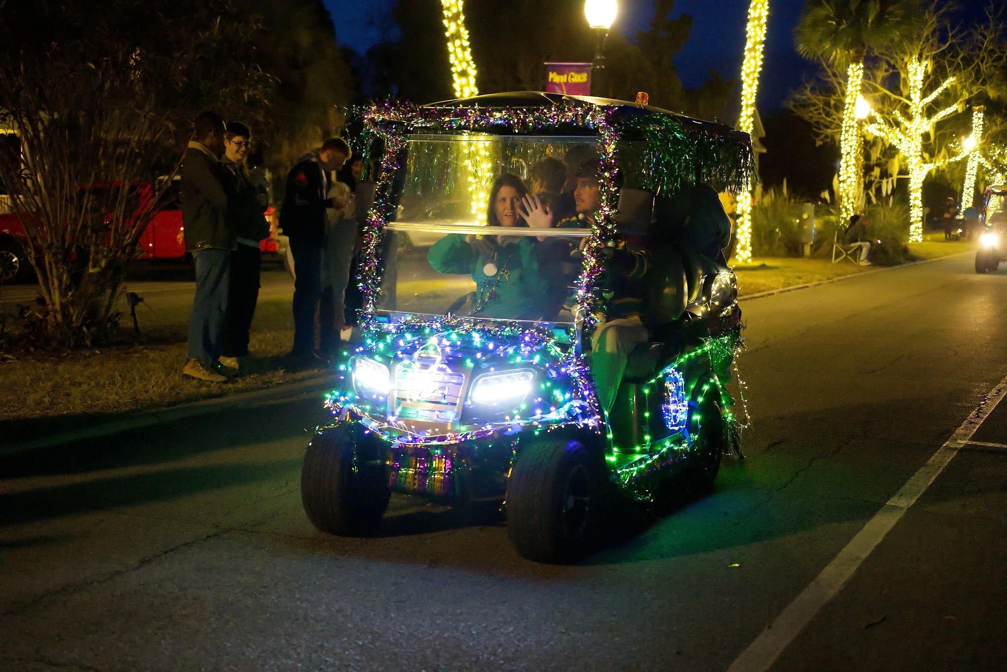 A decorated electric cart with colorful Christmas lights and tinsel, driving along a street at night while a woman waves from the front seat. Several people stand nearby on the sidewalk, and trees wrapped with yellow lights are visible in the backgro