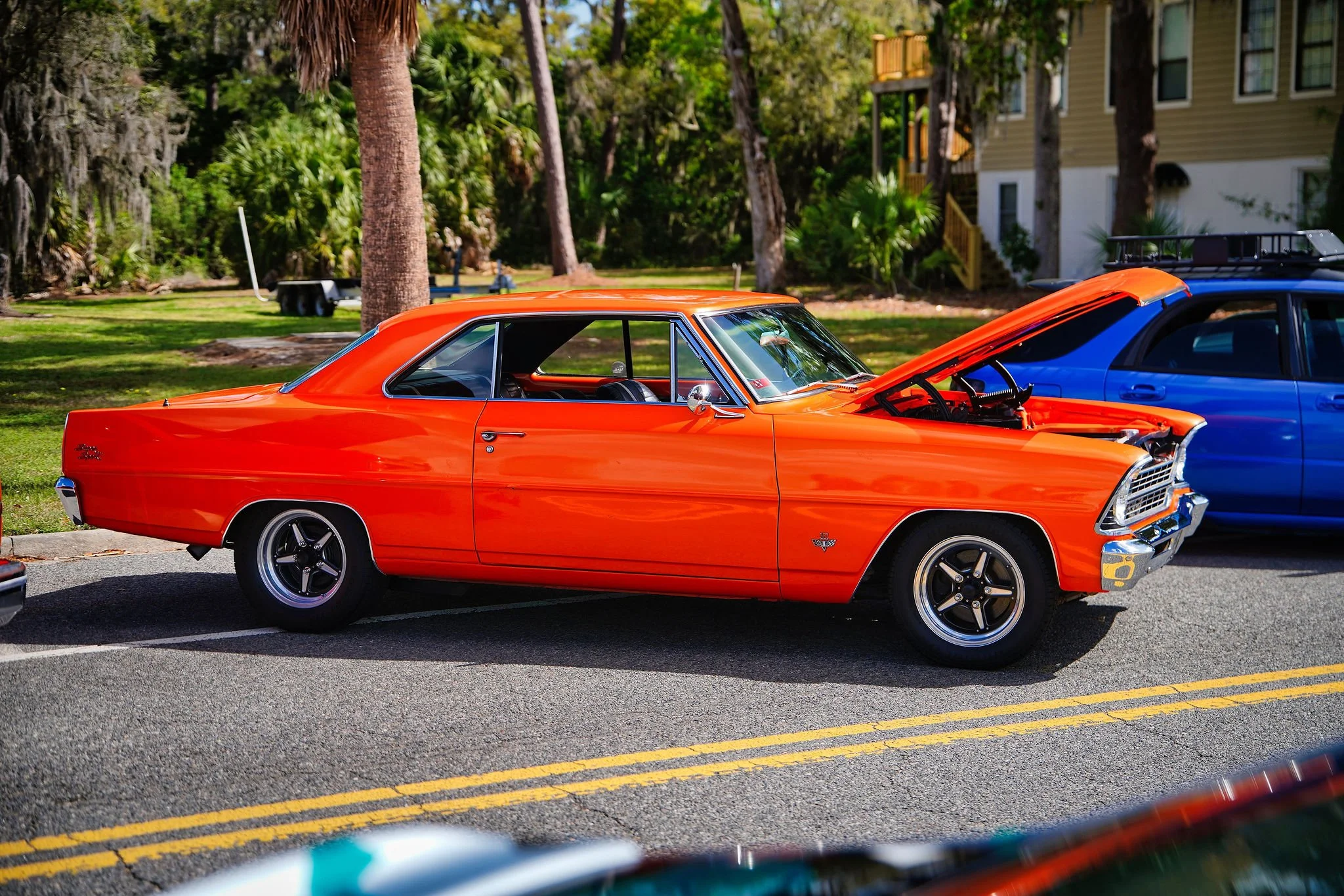 Orange vintage muscle car parked on street with its hood open, next to a blue vehicle, with trees and residential buildings in the background.