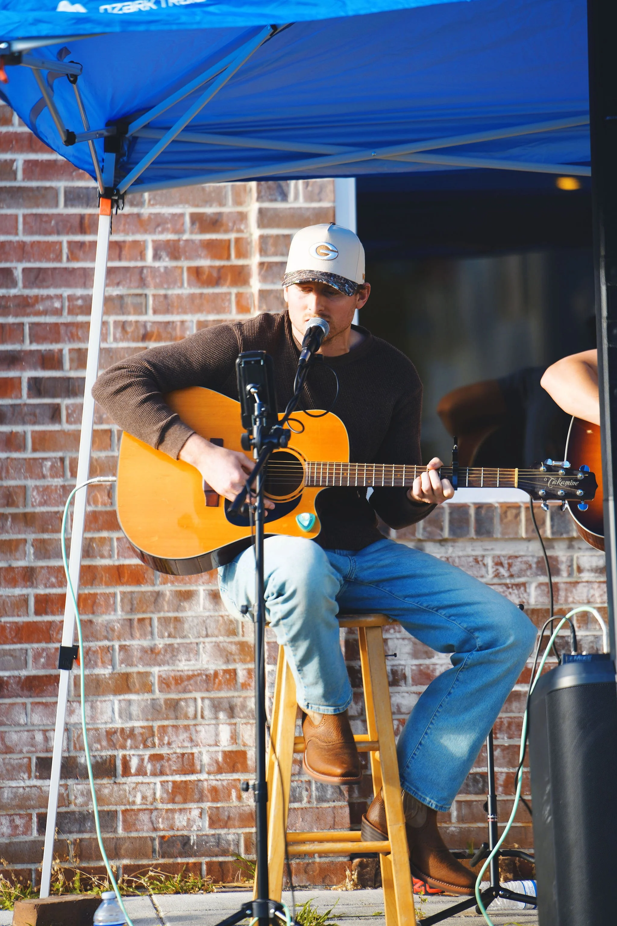 A man wearing a white baseball cap, dark sweater, and jeans plays an acoustic guitar while sitting on a wooden stool and singing into a microphone under a blue canopy, with a brick wall background.