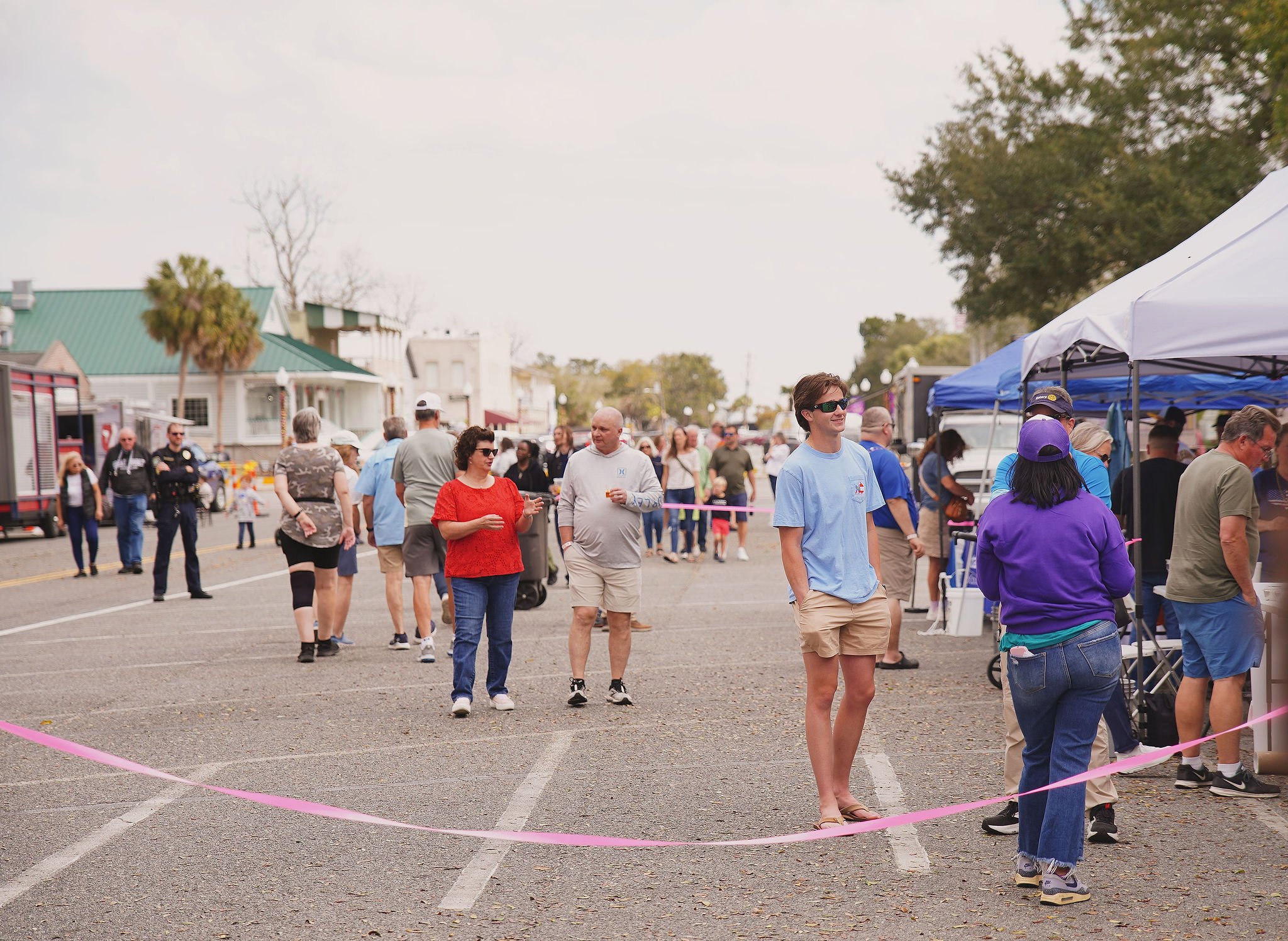 People walking and standing in line at an outdoor event with tents and buildings in the background.