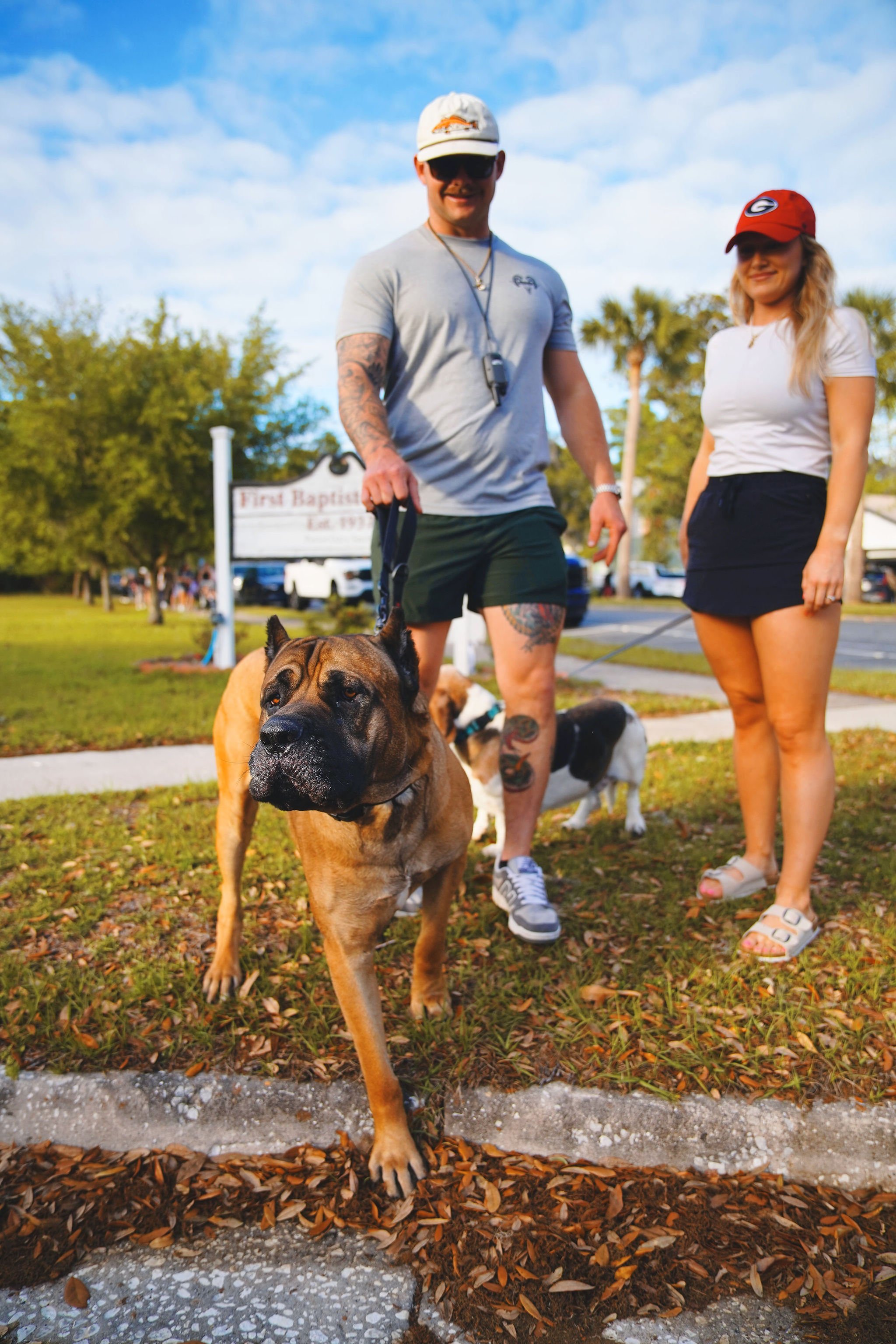 A man and woman standing outside with three dogs near a church. The man wears sunglasses, a hat, and a gray t-shirt, holding a dog's leash. The woman wears a red cap, white t-shirt, and shorts. The brown dog in the foreground is walking toward the ca
