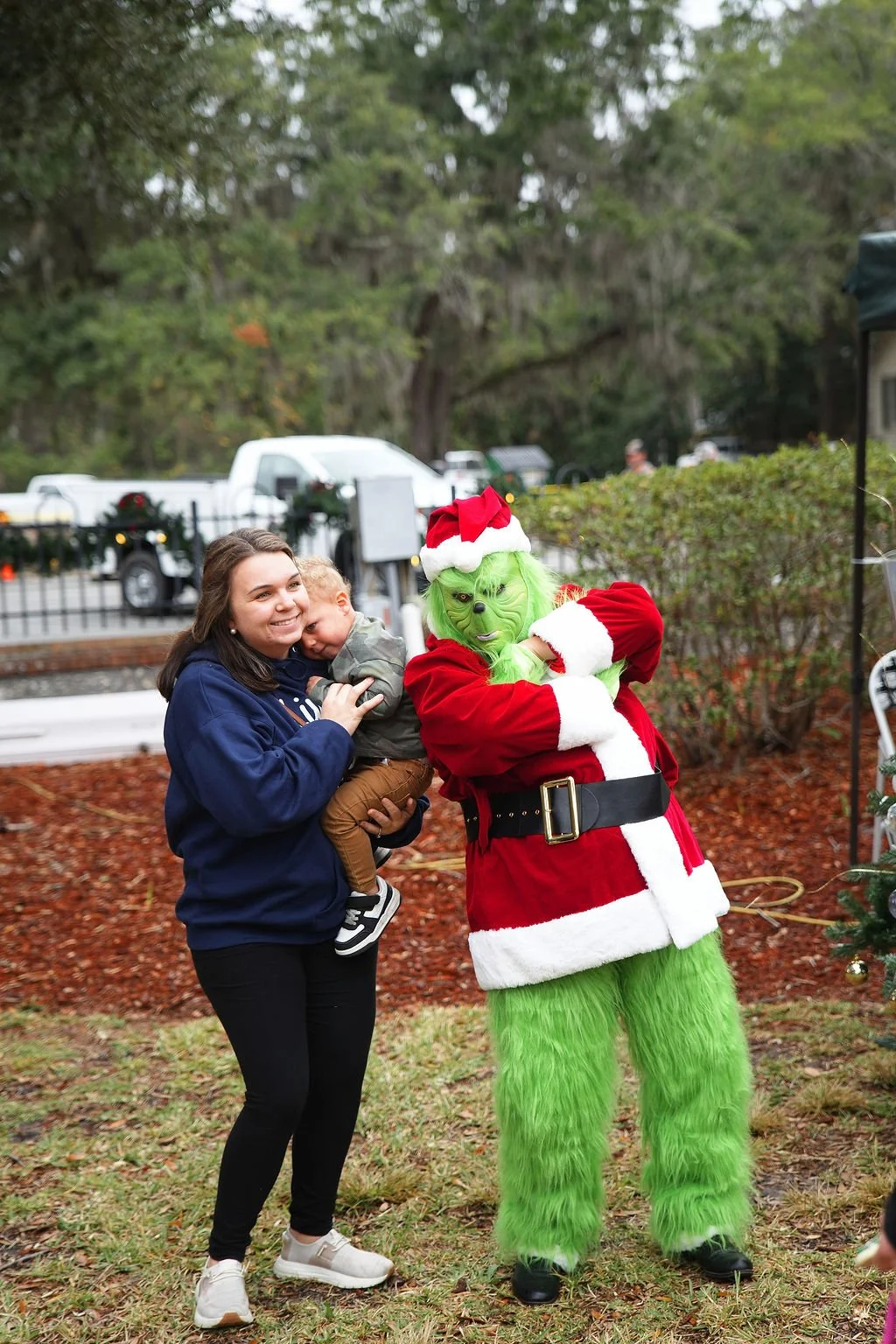 A young woman holding a small child, both smiling, standing outdoors near Christmas decorations and a person dressed as the Grinch in a Santa outfit.