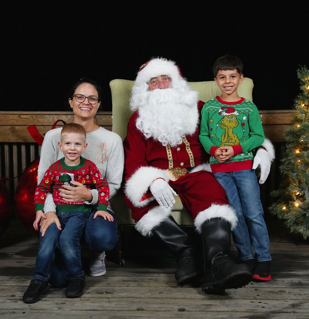 A group of four people with Santa Claus, including two young boys and a woman, all smiling and wearing festive Christmas sweaters, posing indoors with Christmas decorations.