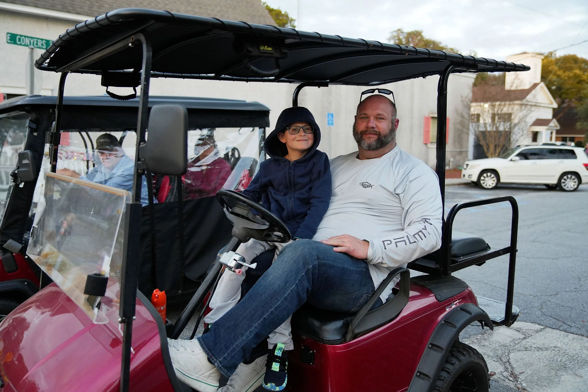 A man and a boy sitting in a golf cart on a street, smiling, with the boy wearing a hooded jacket and the man in a white shirt, surrounded by parked cars and houses.