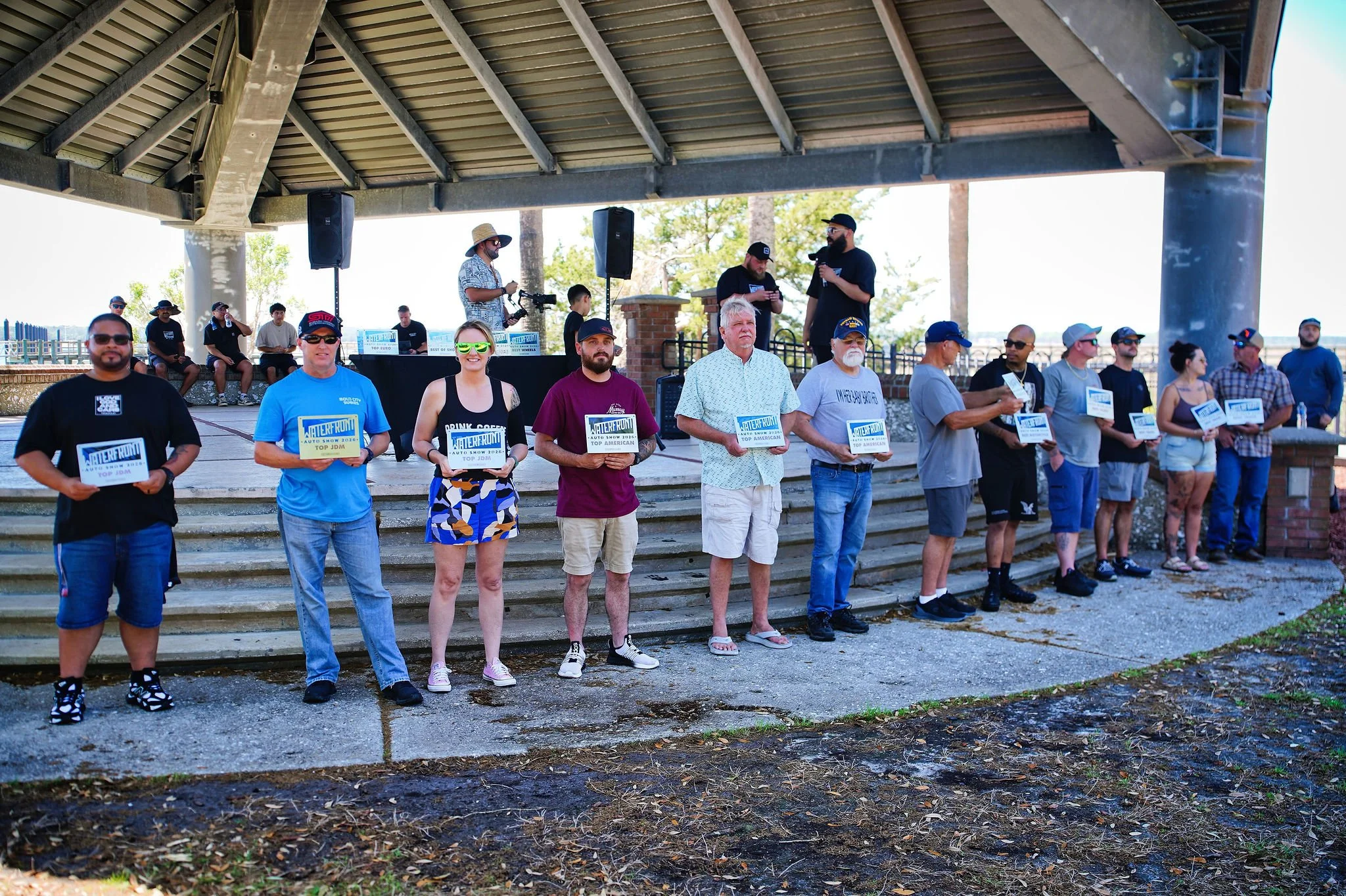 A group of people standing on steps under a pavilion during an outdoor event, holding signs that say 'Waterfront Auto Show 2022 Top American,' with some individuals wearing casual summer clothing, and a few people in the background with microphones a