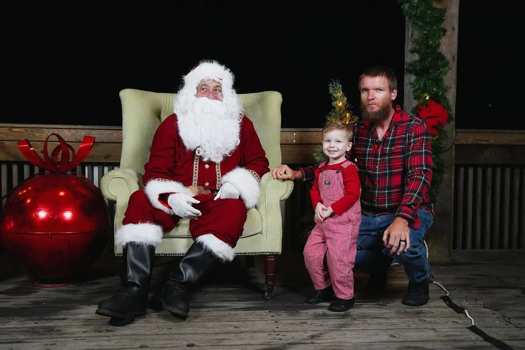 A man dressed as Santa Claus sitting on a green armchair, with a young girl and a man kneeling beside him, all smiling. The girl is wearing red and pink overalls, and the man is in a red plaid shirt. There is Christmas decoration including a large re