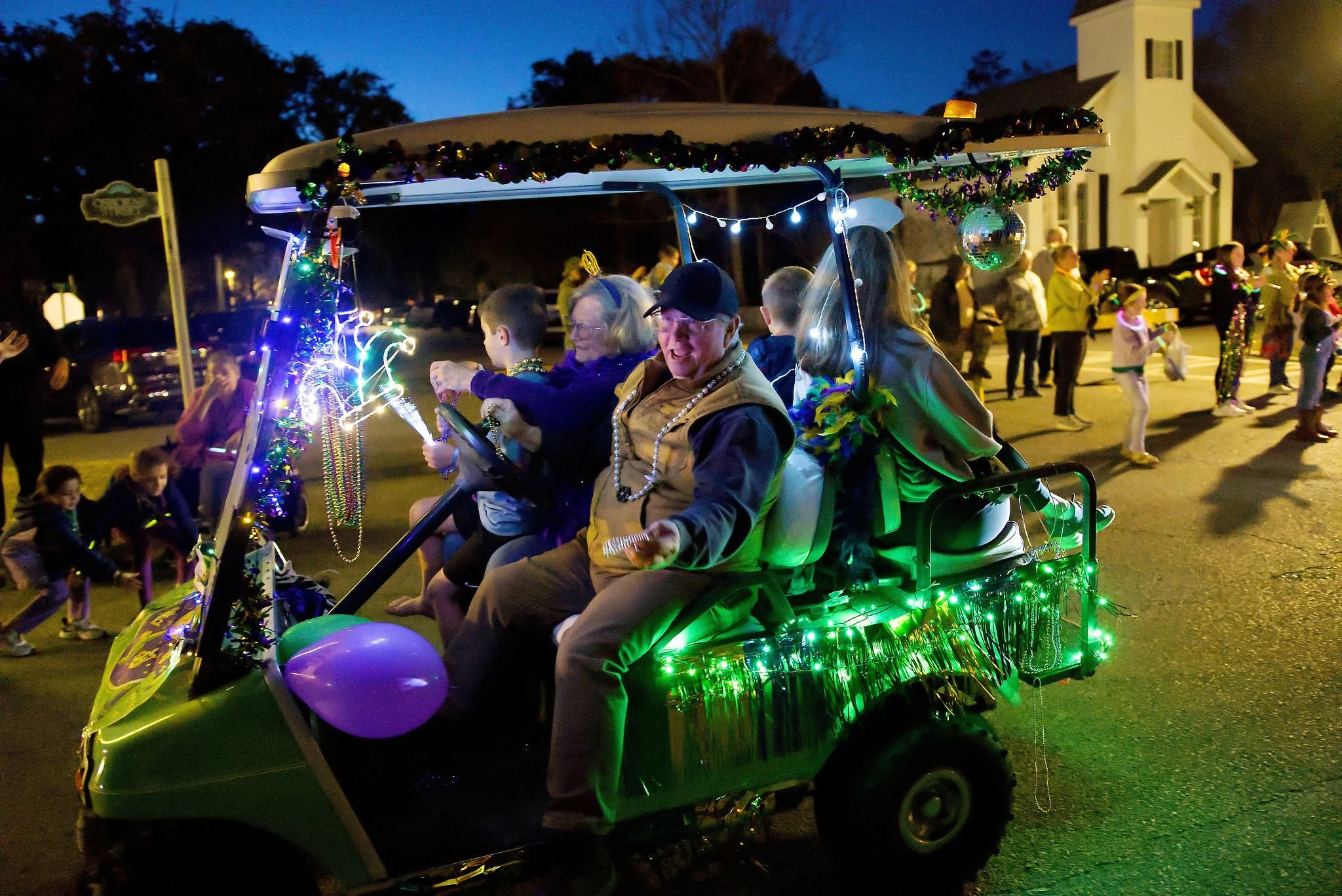 People riding a decorated golf cart during a nighttime parade on a street, with onlookers and children watching.