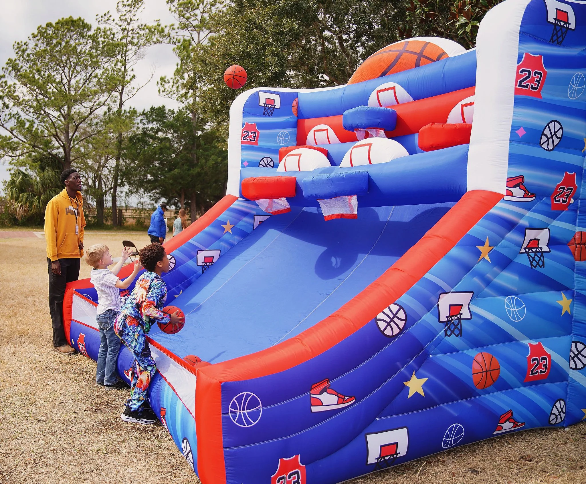 Children playing on a basketball-themed inflatable slide outdoors on a grassy field, supervised by an adult, with trees and cloudy sky in the background.