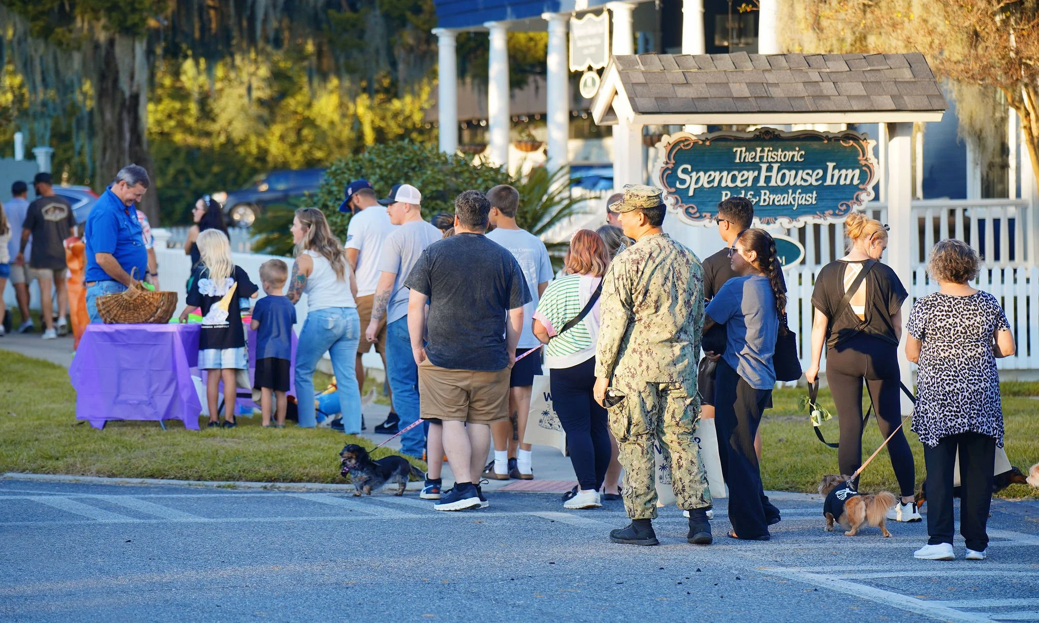 A group of people waiting in line outside a building with a sign that reads "The Historic Spencer House Inn Bed & Breakfast." The line includes adults, children, and a woman in military uniform, with some people holding small dogs.