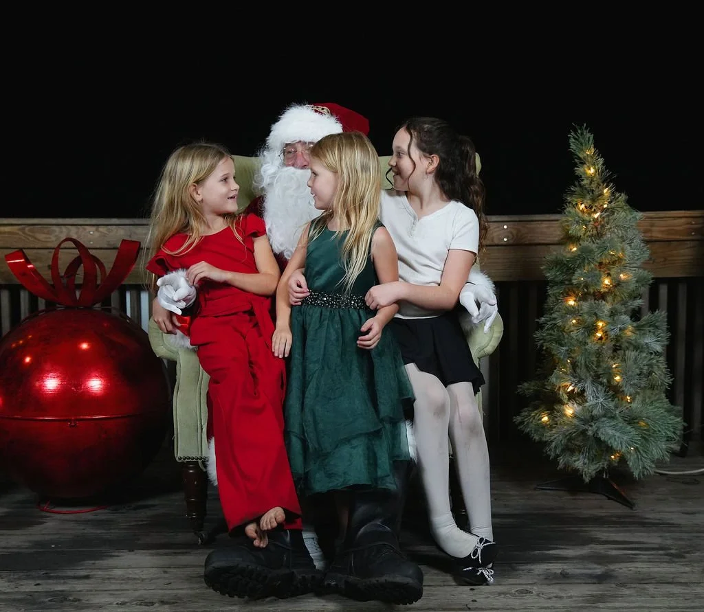 Three children sitting with Santa Claus on a green chair, surrounded by Christmas decorations including a large red ornament and a small decorated Christmas tree, indoors with a black background.
