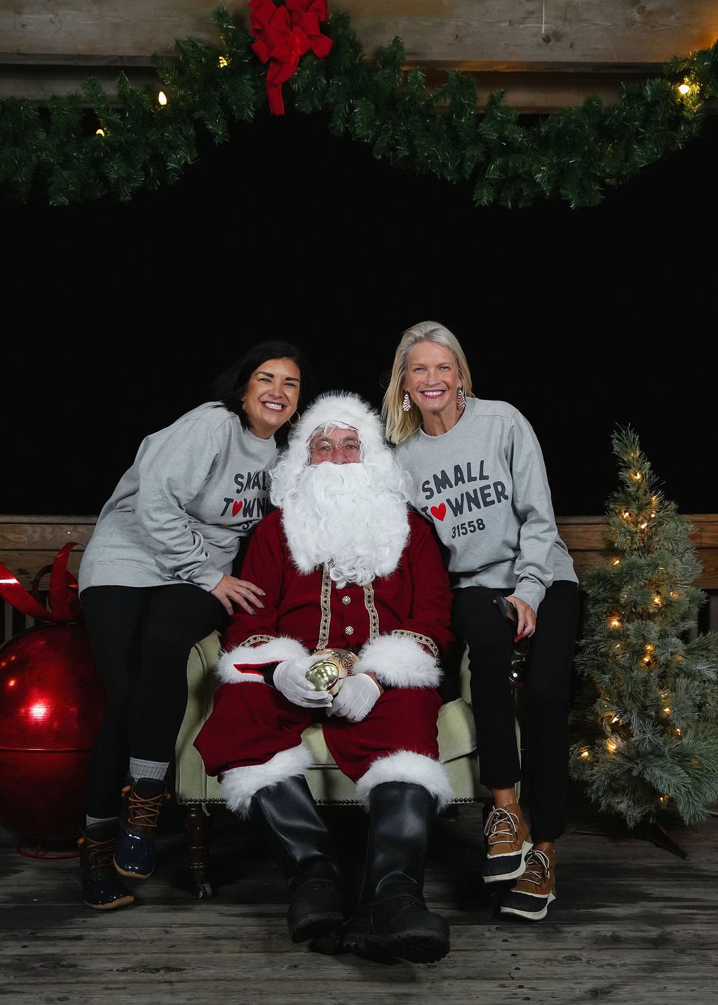 Two women and a man dressed as Santa Claus sitting on a bench with Christmas decorations, including a small Christmas tree and large ornaments, in what appears to be a holiday event setting.
