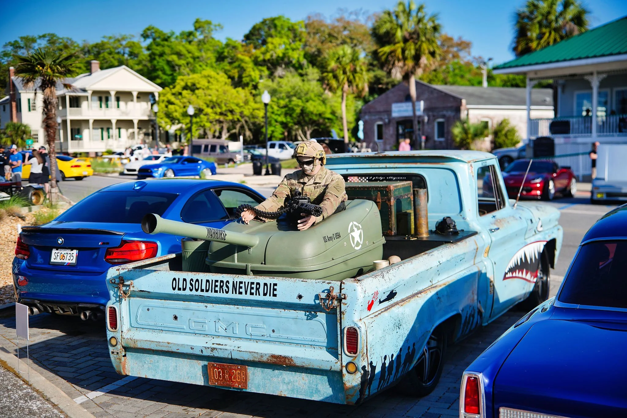 A vintage GMC truck with a painted shark face on the side, displaying the words 'Old Soldiers Never Die.' A soldier mannequin dressed in military gear is seated in the truck's bed, holding a gun. The truck is parked on a street with other cars and pe