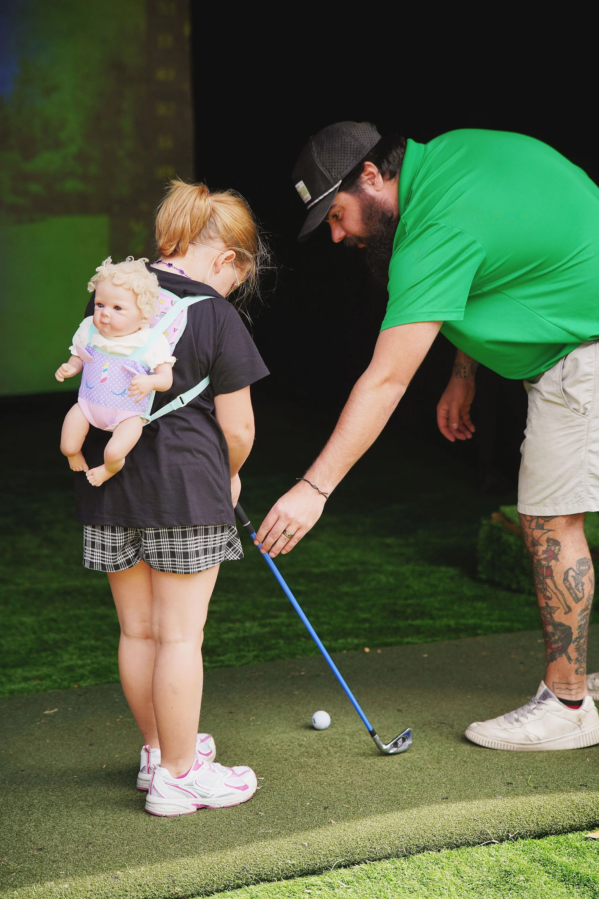 A girl with a doll on her back learning to golf with an instructor helping her.