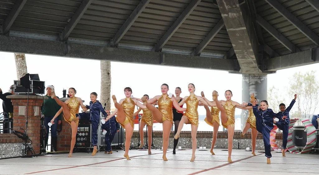 Group of young dancers practicing under a pavilion, wearing gold and navy costumes, some in gold dresses and others in navy tracksuits, during an outdoor dance rehearsal.