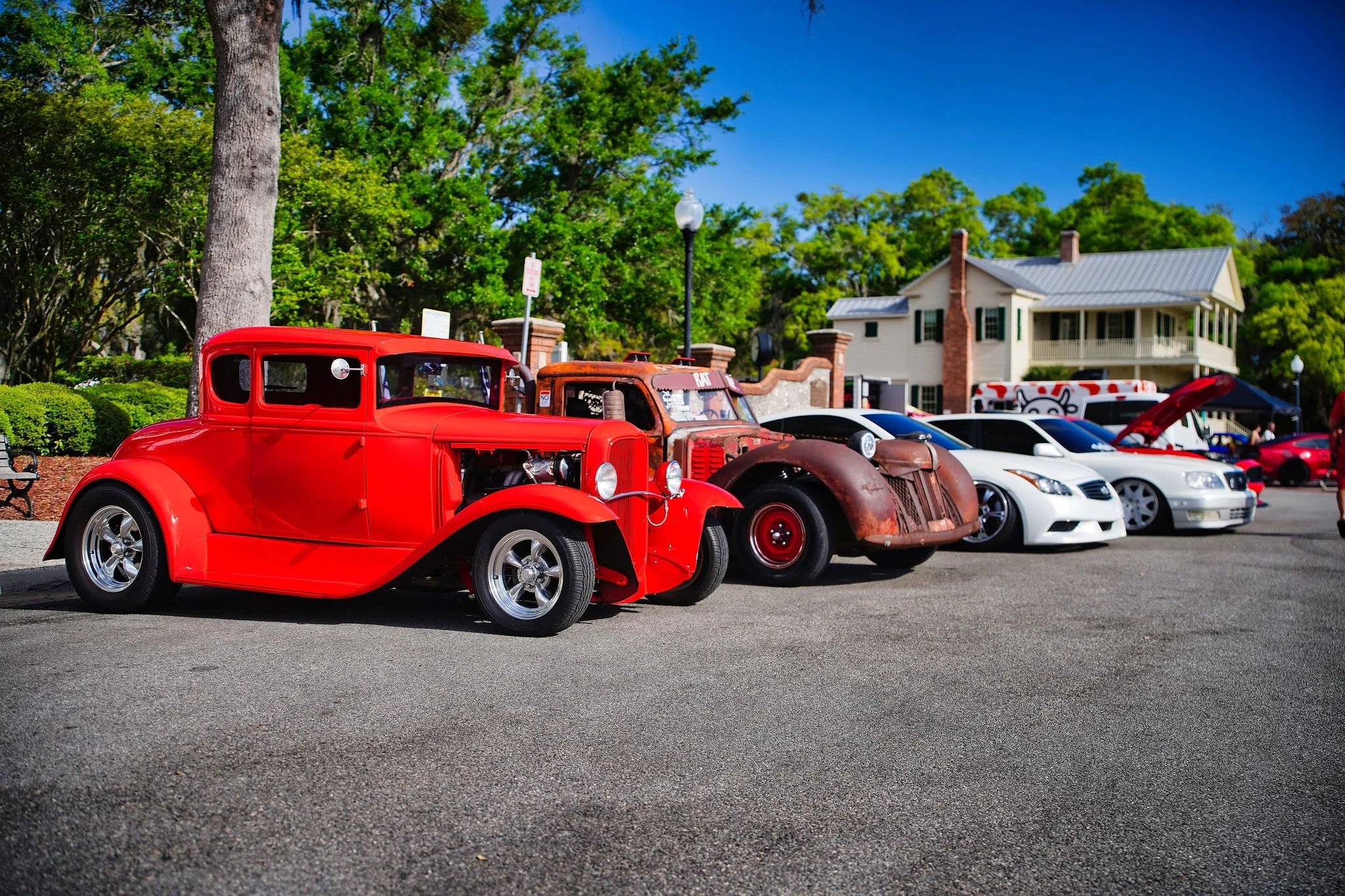 Line of vintage and modern cars parked on street during a car show, with trees and a house in the background.