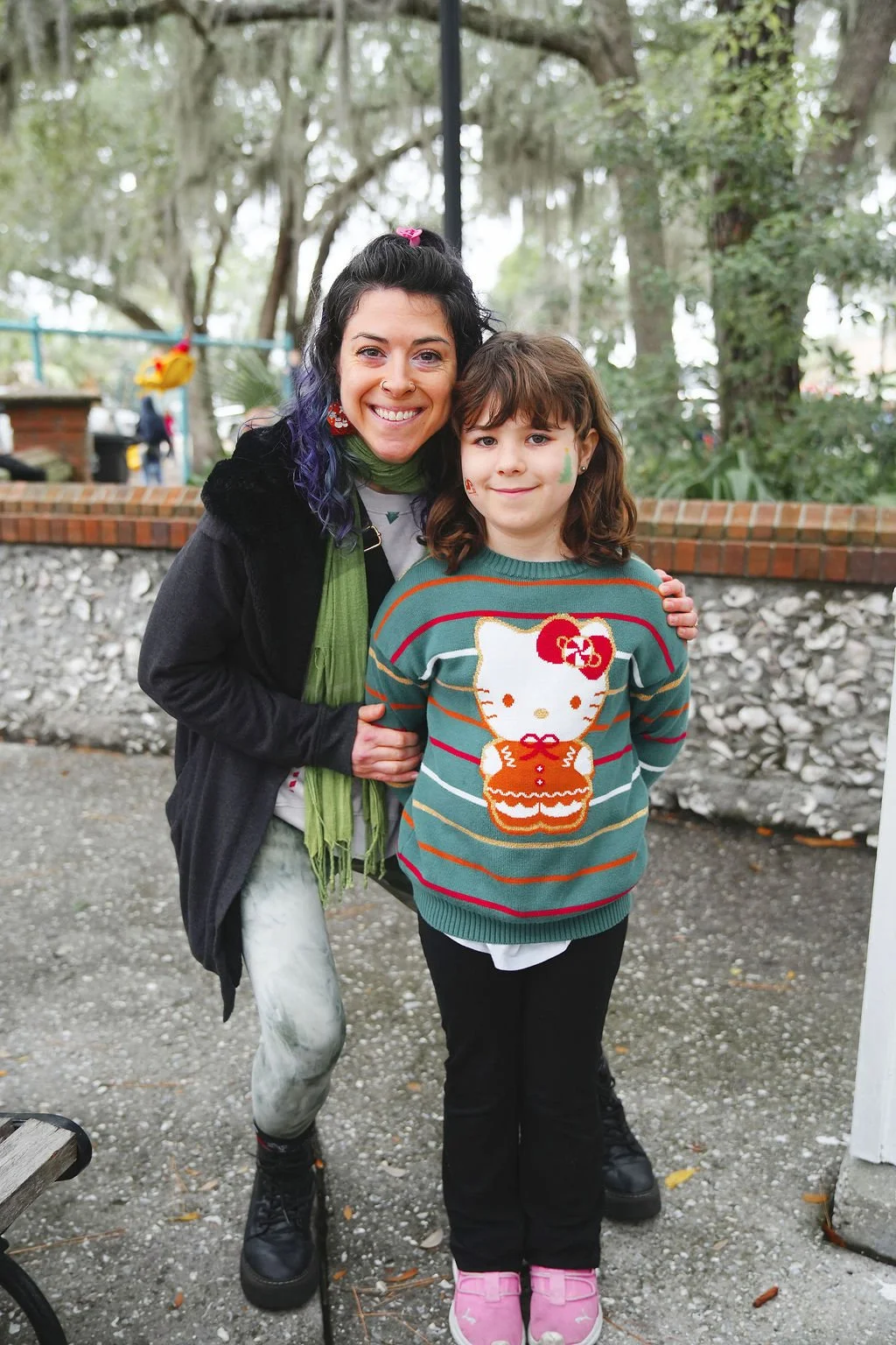 A woman and a young girl smiling and posing outdoors. The woman has dark hair with purple tips, wearing a black hoodie, a green scarf, and jeans. The girl has brown hair with face paint on her cheeks, wearing a striped sweater with Hello Kitty, and p